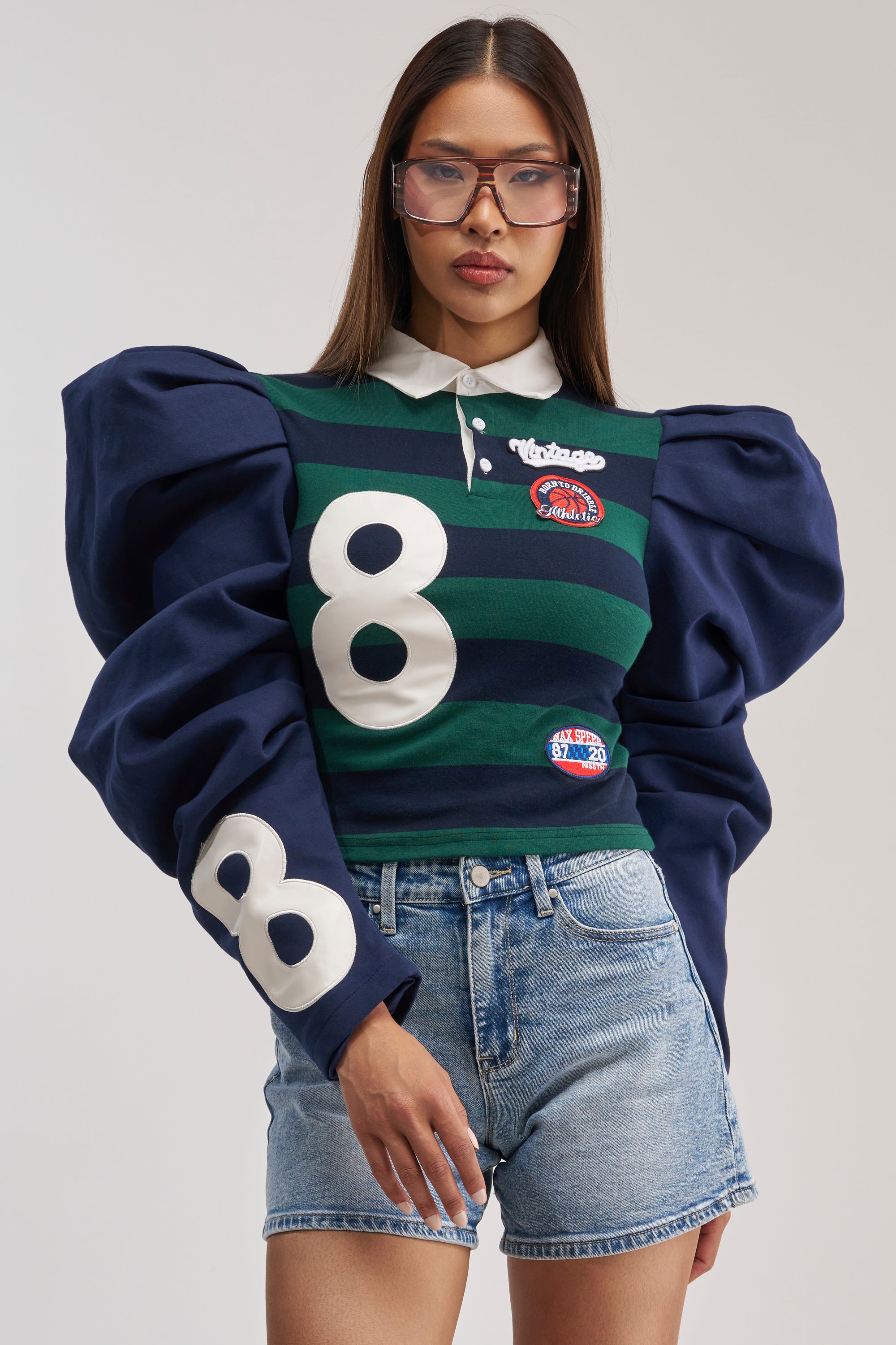 A woman models the COLLEGE DAYS STRIPE POLO, featuring oversized puff sleeves, bold stripes, and prominent varsity number 8 patches. She pairs it with denim shorts and tinted sunglasses while standing against a plain background.