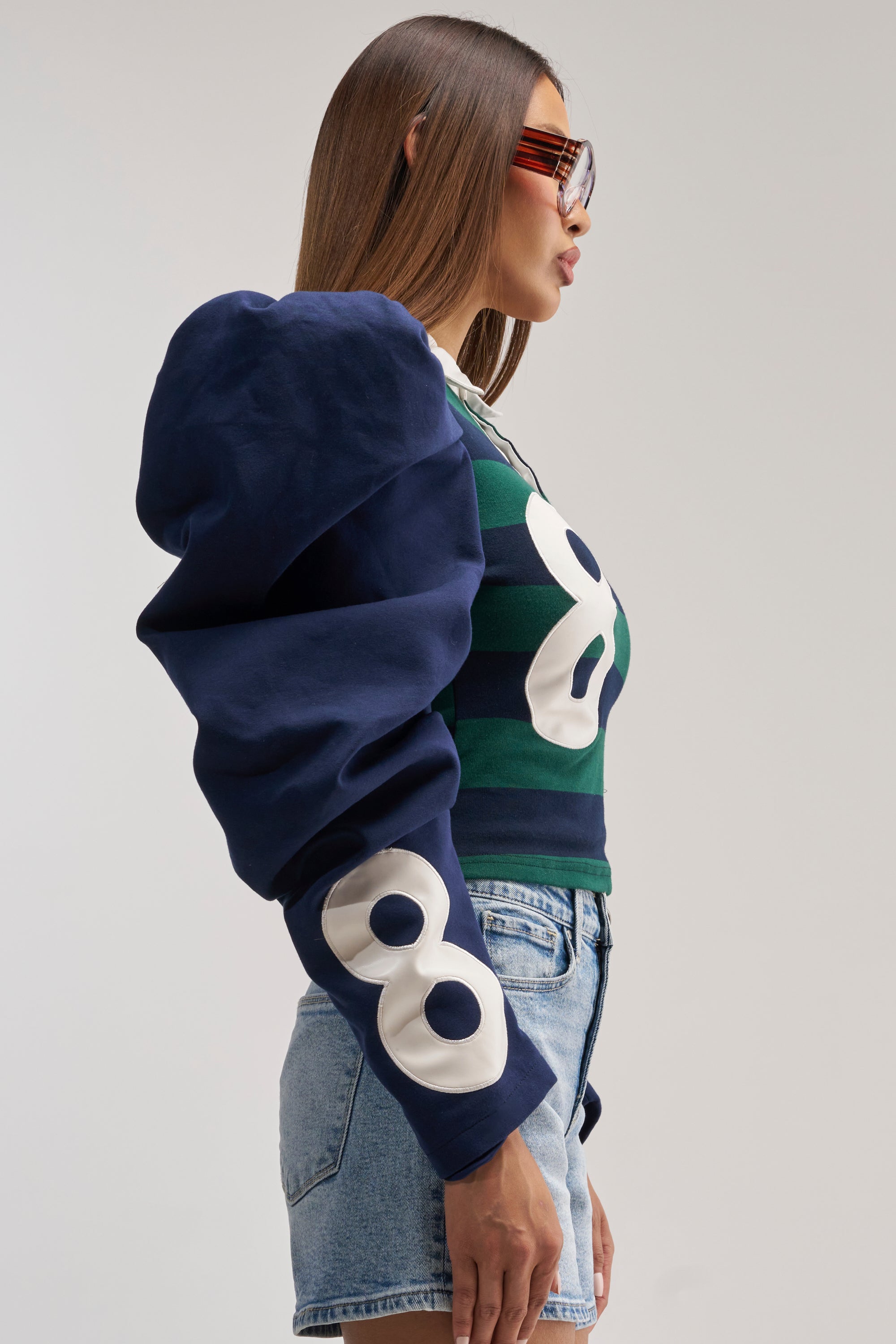 A woman in sunglasses stands in profile against a plain background, wearing the COLLEGE DAYS STRIPE POLO—a blue and green striped polo shirt—paired with blue shorts and a navy varsity jacket featuring large white "8" patches on the sleeve.