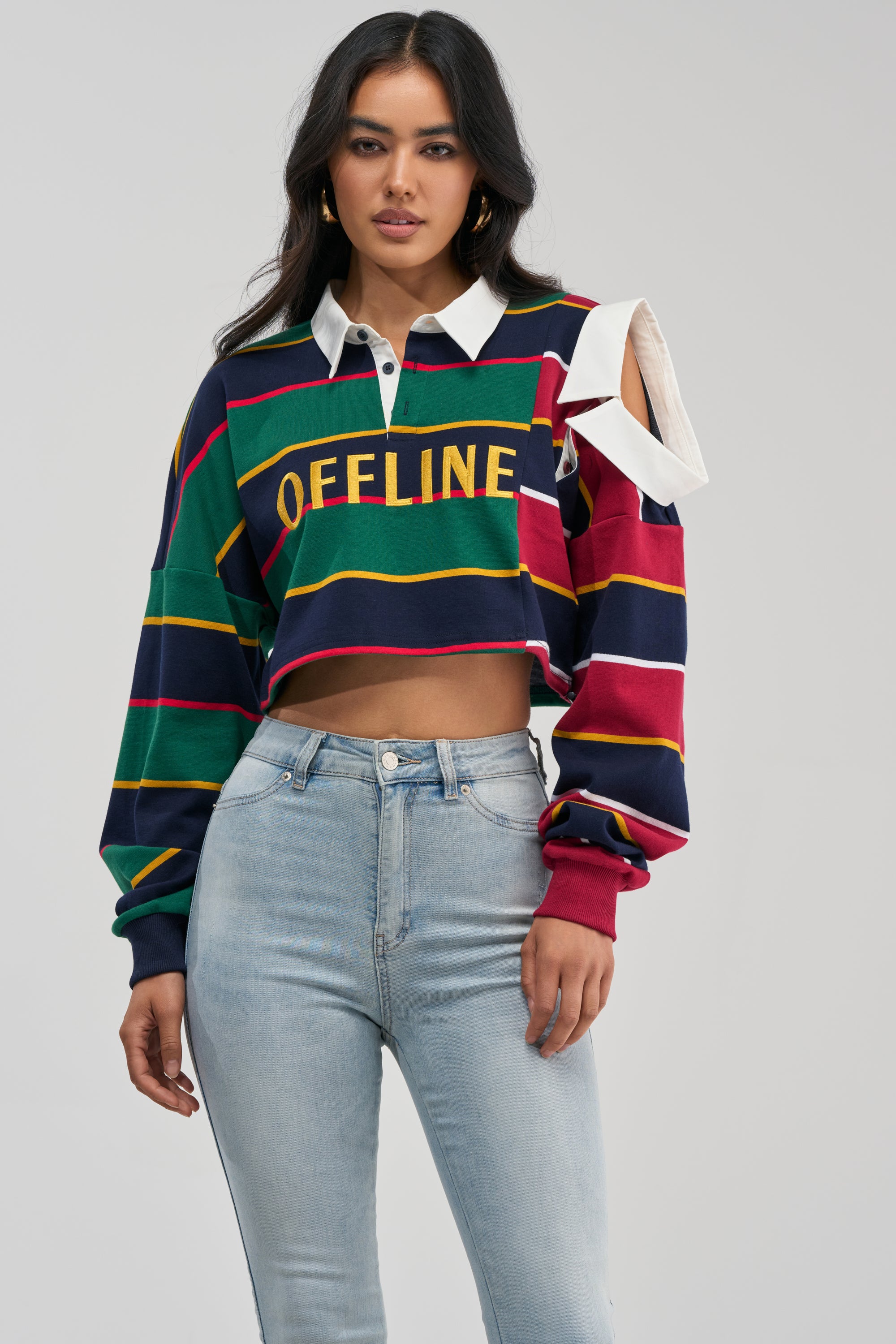 A woman stands against a plain background, styled in light blue jeans and the OFFLINE CROPPED POLO SHIRT—a multicolored, striped rugby shirt with a cut-out detail on one shoulder, evoking a school girl streetwear vibe.