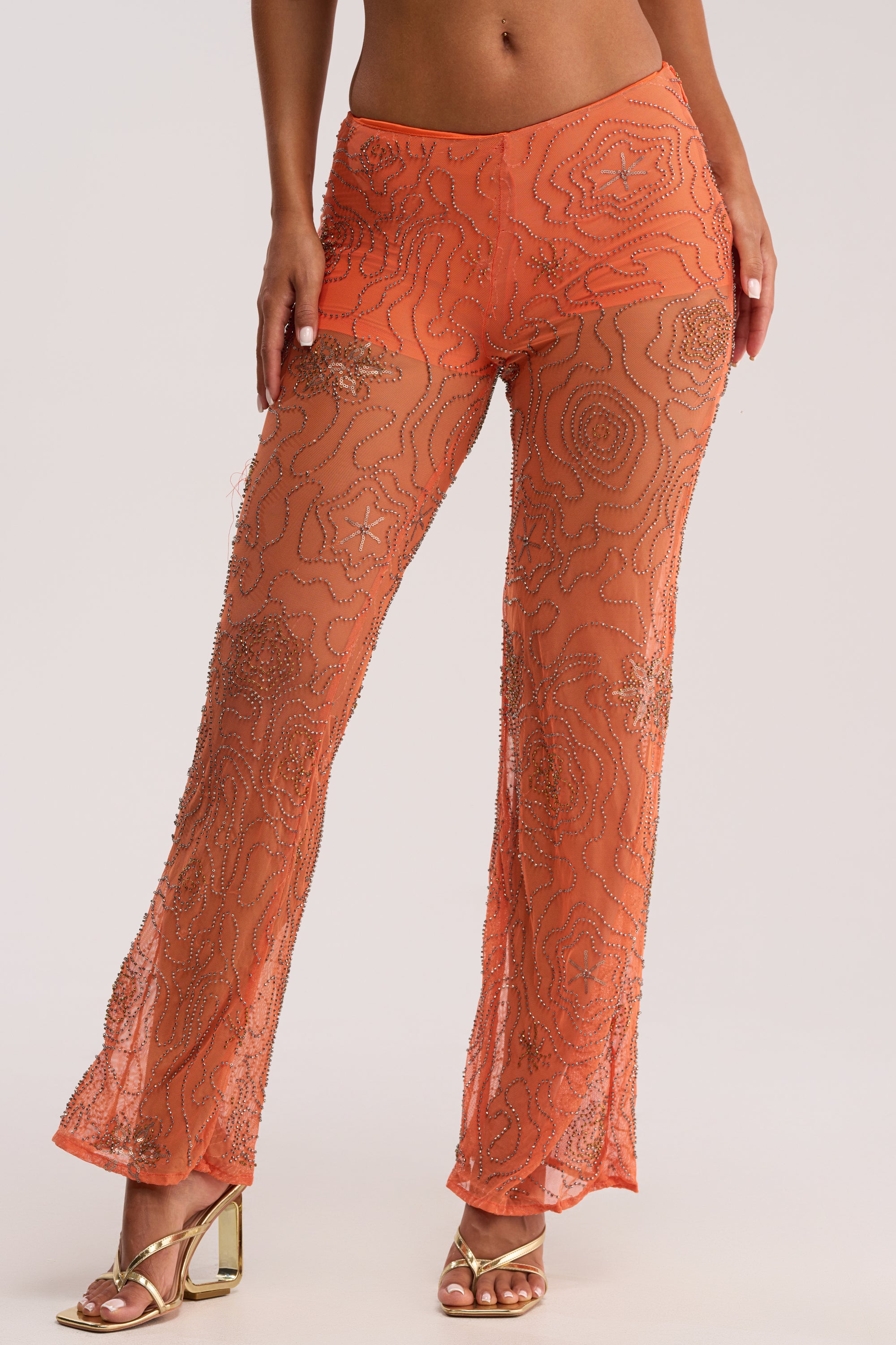 A person wearing the KATE EMBELLISHED MESH PANT IN ORANGE, featuring sheer fabric and intricate beadwork, paired with gold high-heeled sandals.