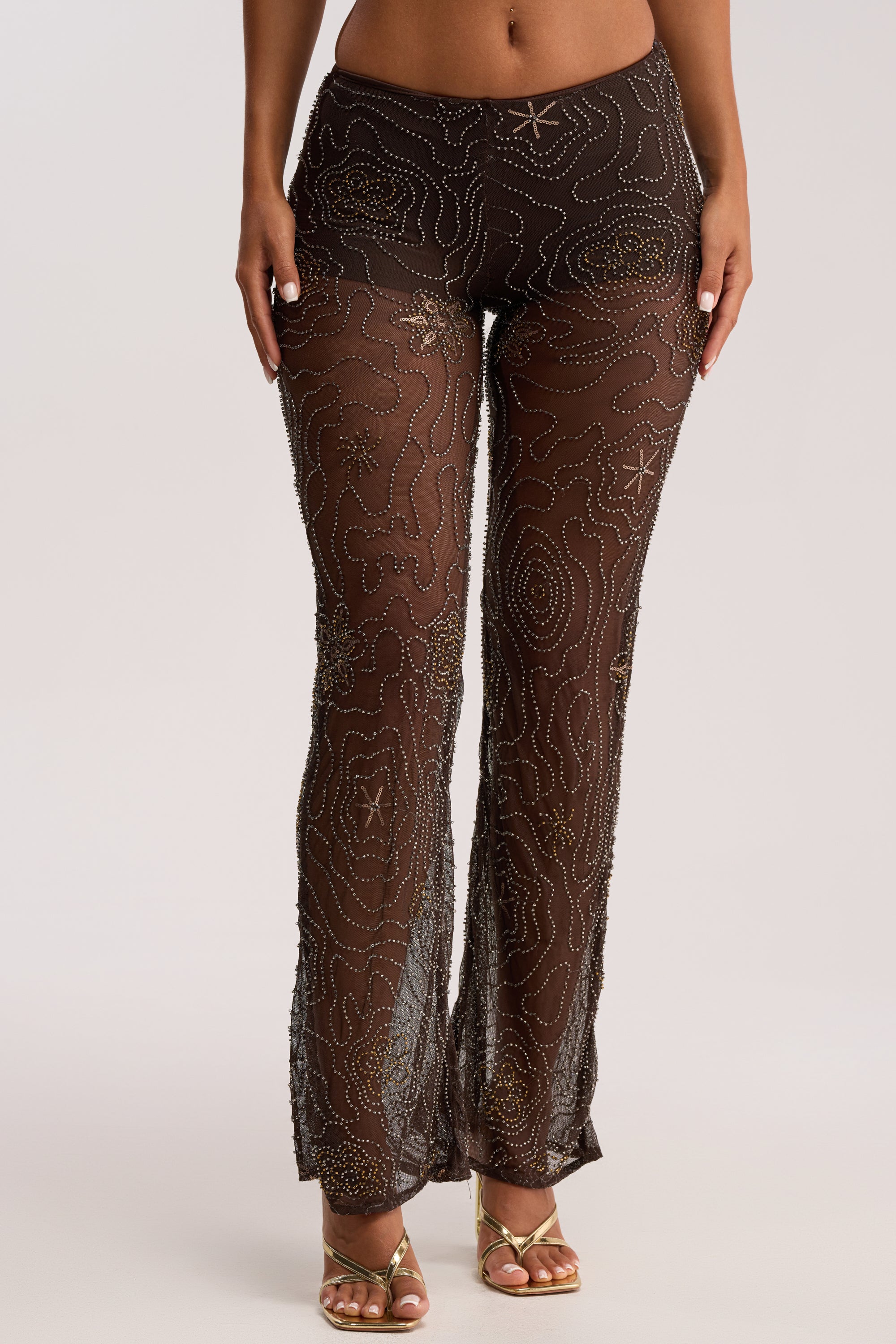 A person wears the KATE EMBELLISHED MESH PANT in sheer brown, featuring high waist, metallic bead mesh with swirling and star patterns, styled with gold strappy heels against a plain neutral background.