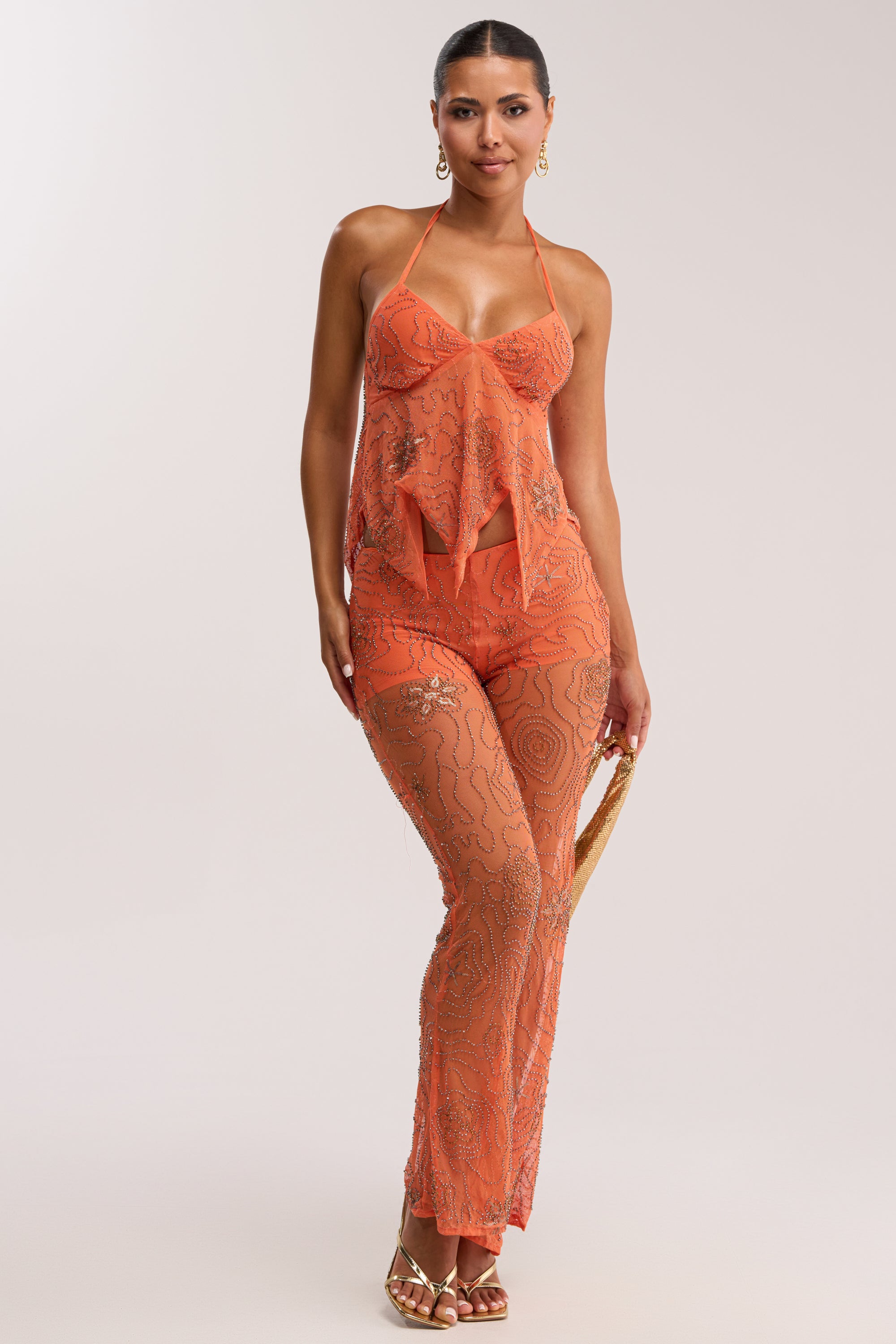 Woman wears the KATE EMBELLISHED MESH TOP IN ORANGE with matching flared pants, confidently posing with one hand on her hip, gold hoop earrings, and heels against a plain light background.