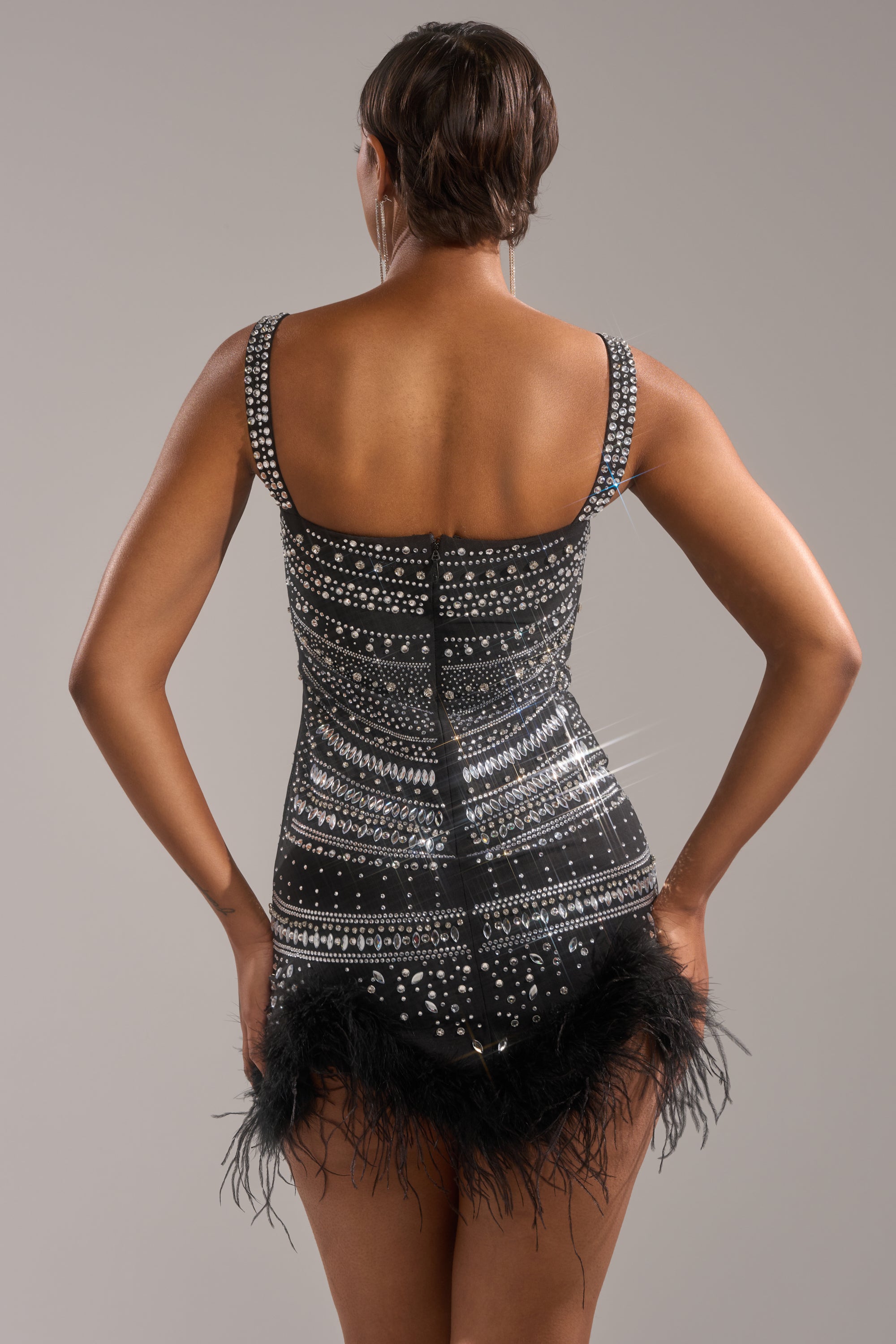 A person faces away wearing the SHOWSTOPPER RHINESTONE MINI DRESS, a black bodycon mini with feather trim and sparkling silver sequins and beads, set against a plain neutral background.