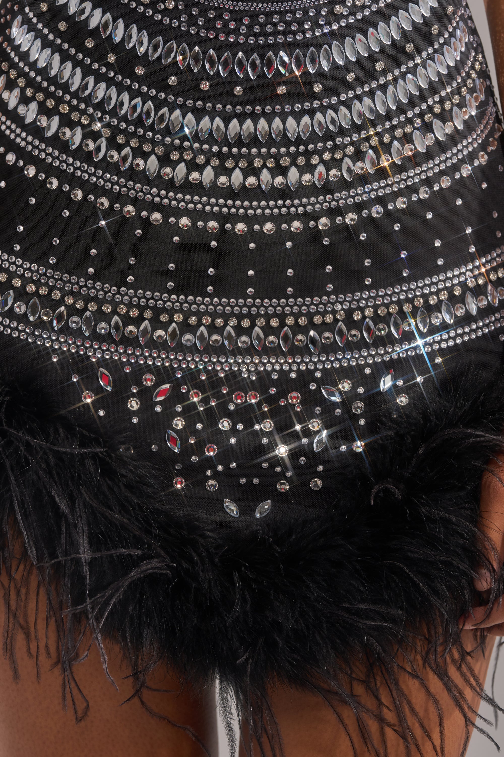 A close-up of the SHOWSTOPPER RHINESTONE MINI DRESS, a bodycon LBD adorned with silver rhinestones and sequins, featuring a black feather-trimmed hem. Two hands by the sides highlight this chic feather trim dress.