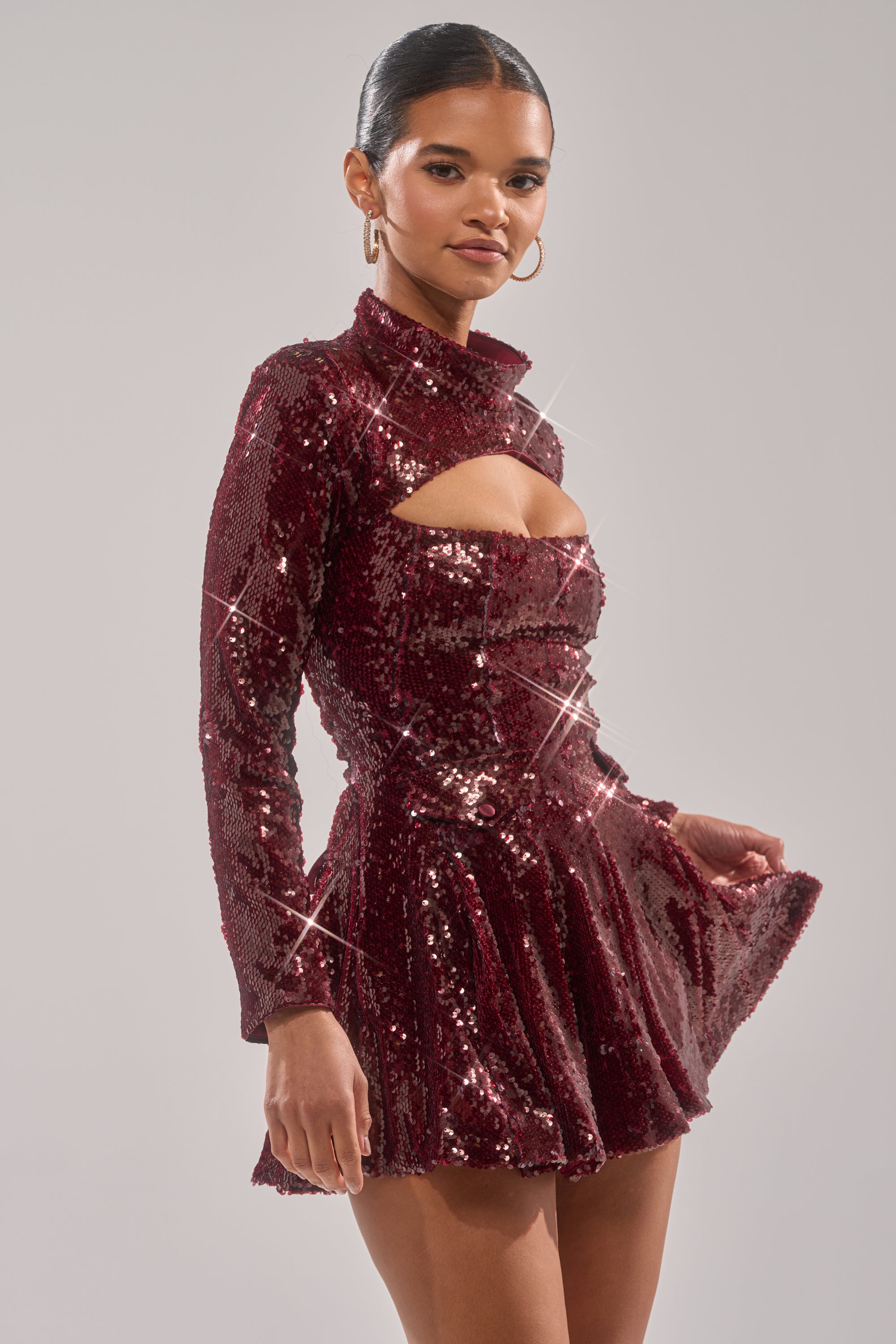 A woman models the IM YOUR FAVORITE SEQUIN MINI DRESS, a shiny burgundy mini with long sleeves, high neckline, and chest cutout. She pairs it with hoop earrings and slicked-back hair against a plain light gray background.