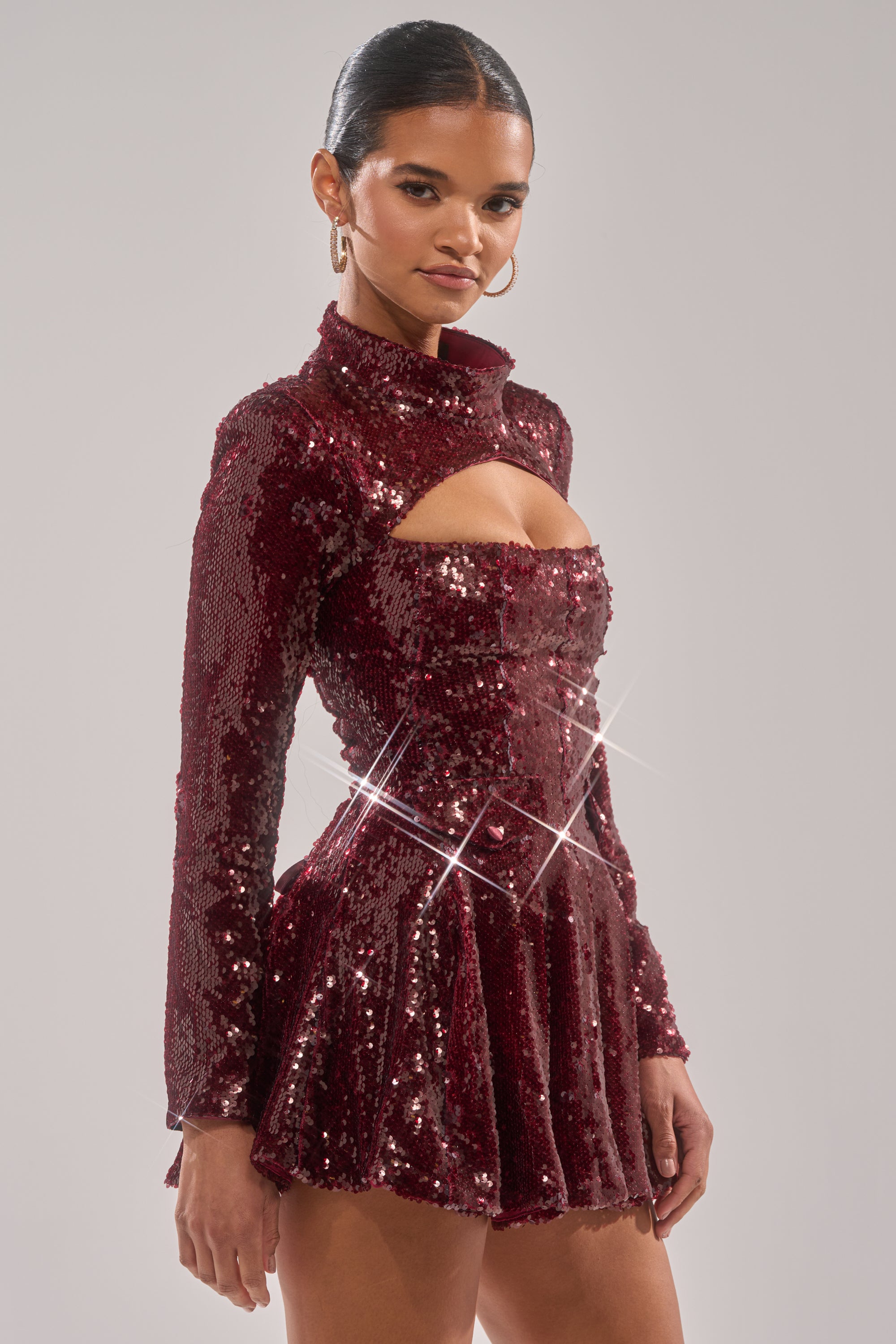 A woman models the IM YOUR FAVORITE SEQUIN MINI DRESS—a sparkly deep red sequin mini with long sleeves, a mock neck, and a chest cut-out. She completes the look with slicked-back hair, hoop earrings, and a confident pose.