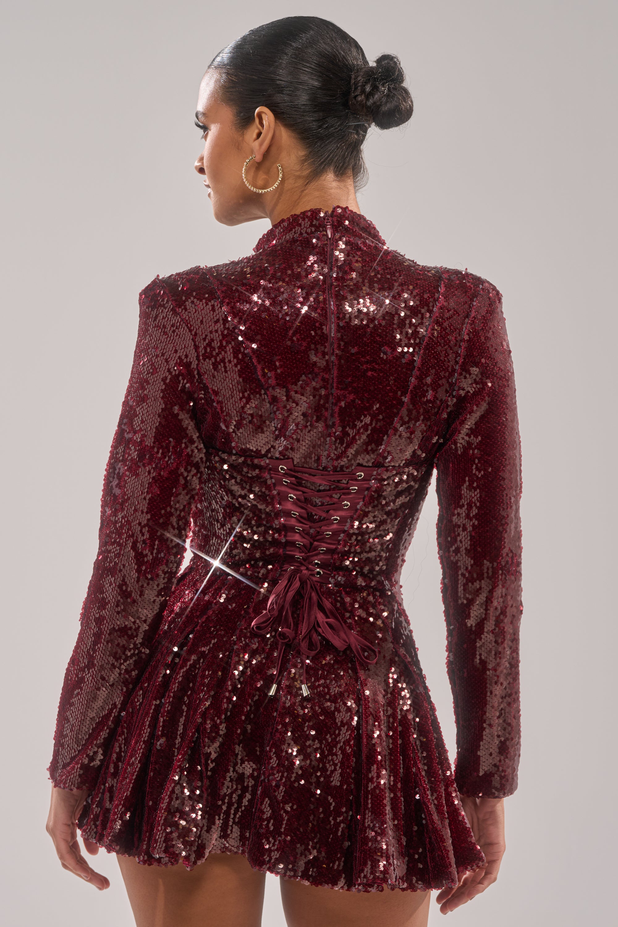 A person with hair in a neat bun wears the IM YOUR FAVORITE SEQUIN MINI DRESS, a long-sleeved, dark red festive dress with sequins, a corset-style lace-up back, and pleated skirt, standing against a plain light gray background.