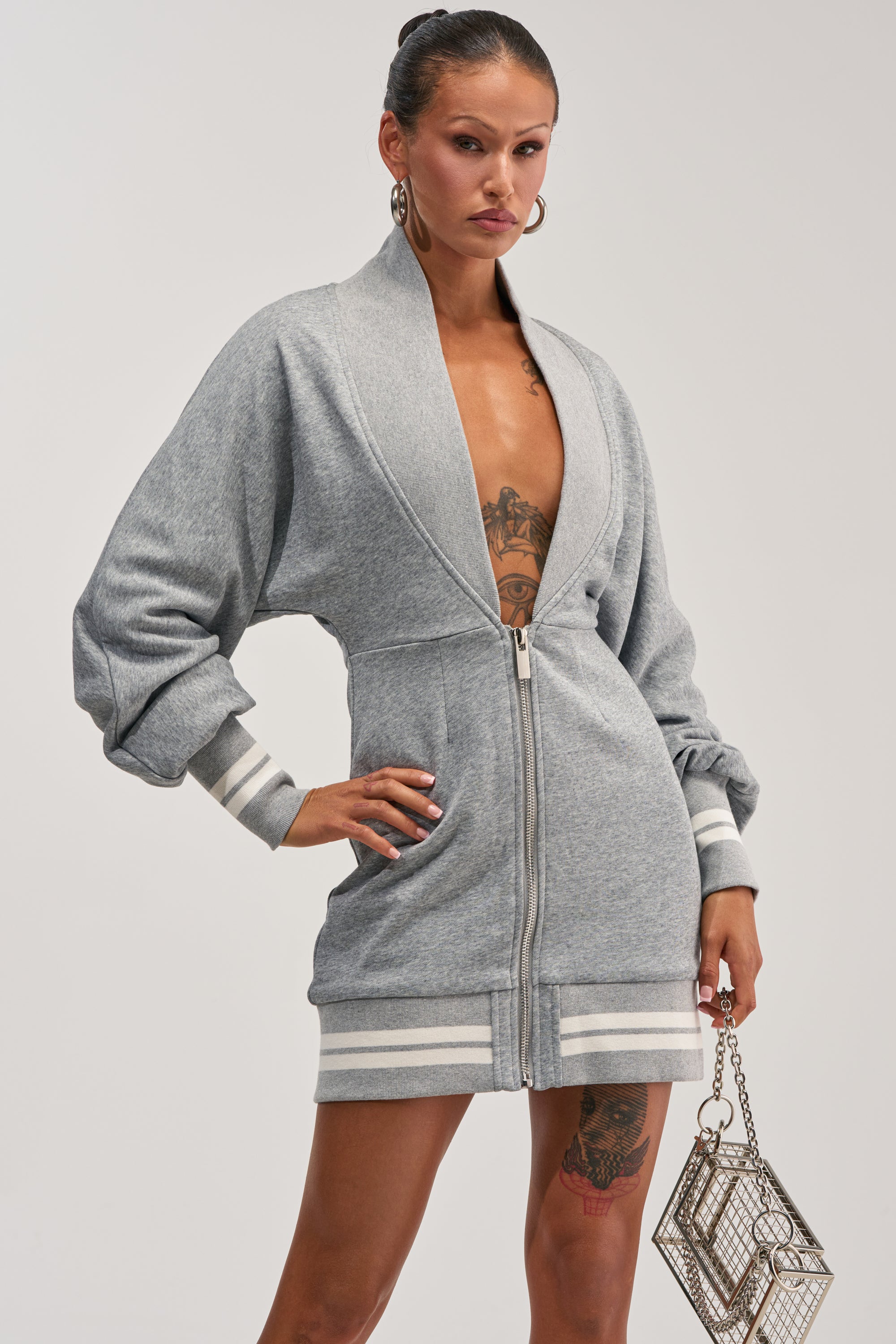 Model wears the FAST LANE MINI DRESS IN HEATHER GREY, a zip-front mini dress with long sleeves and white stripe detailing at the cuffs and hem. She pairs it with large hoop earrings, displays bold tattoos, and accessorizes with a geometric silver handbag for a chic modern streetwear look.