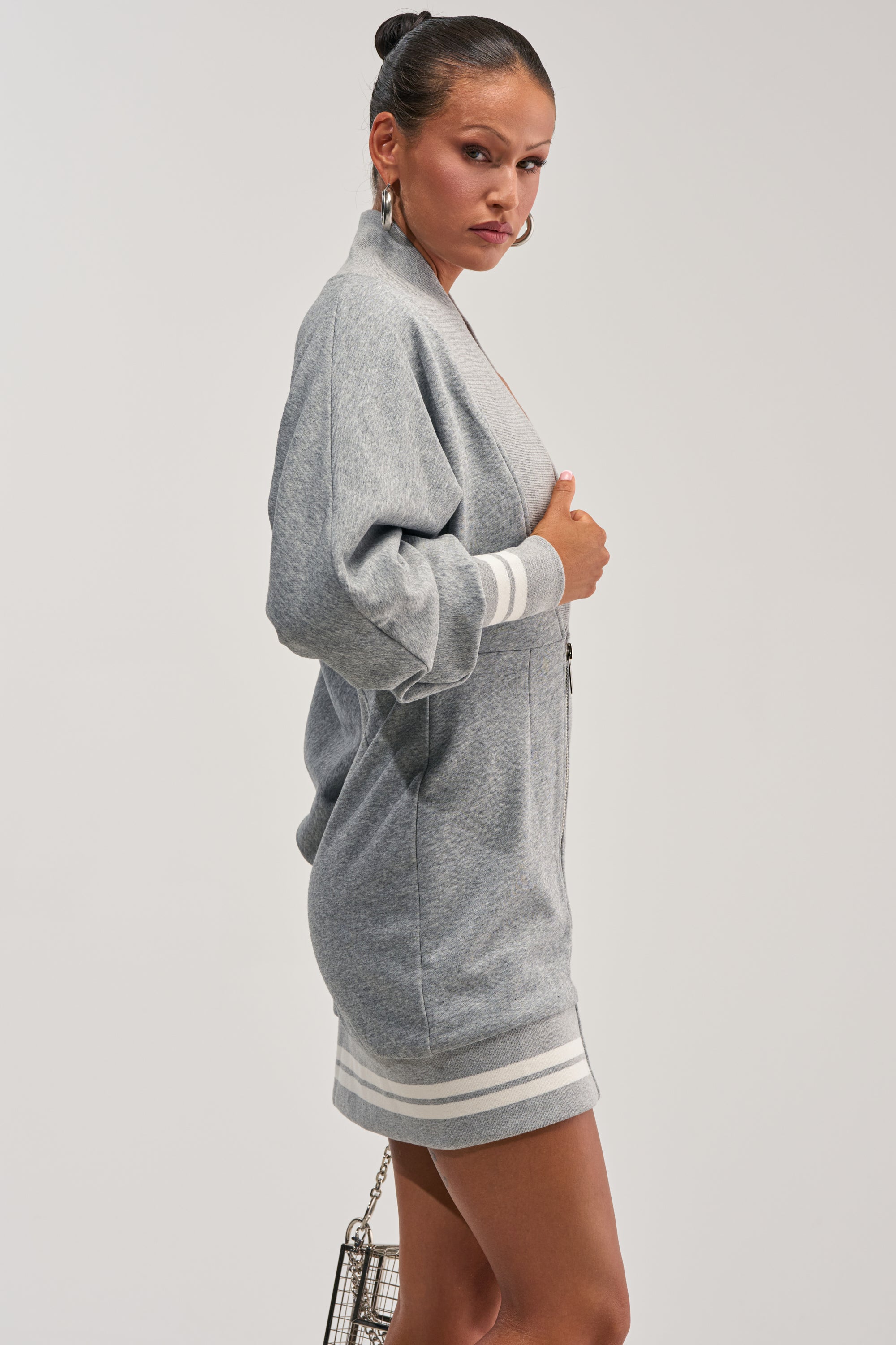 A woman stands sideways against a plain background, looking at the camera while holding a small silver purse and wearing the FAST LANE MINI DRESS IN HEATHER GREY—a zip-front style featuring white racing stripes on the cuffs and hem.