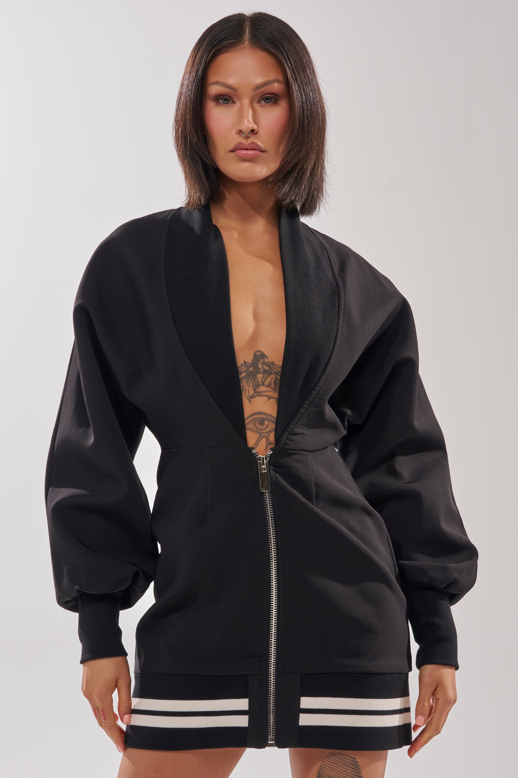 A woman with straight dark hair and visible chest tattoos confidently wears the FAST LANE MINI DRESS—a sporty black mini dress with wide sleeves and varsity-style white stripes—standing against a plain, light background.