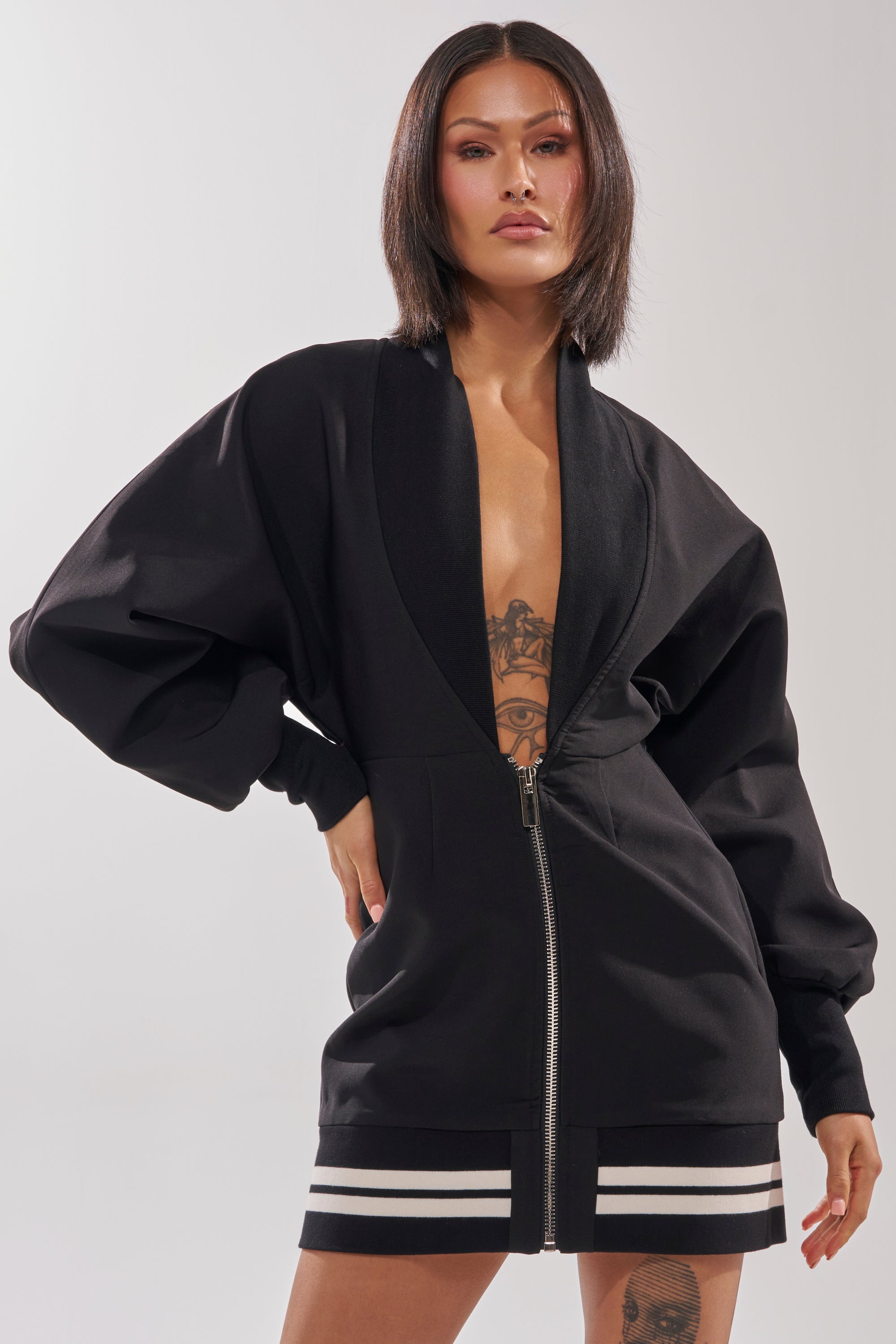A woman with straight dark hair confidently wears the FAST LANE MINI DRESS—a sporty chic black zip-up mini dress with wide sleeves and white stripes near the hem—showing a torso tattoo against a plain light background.
