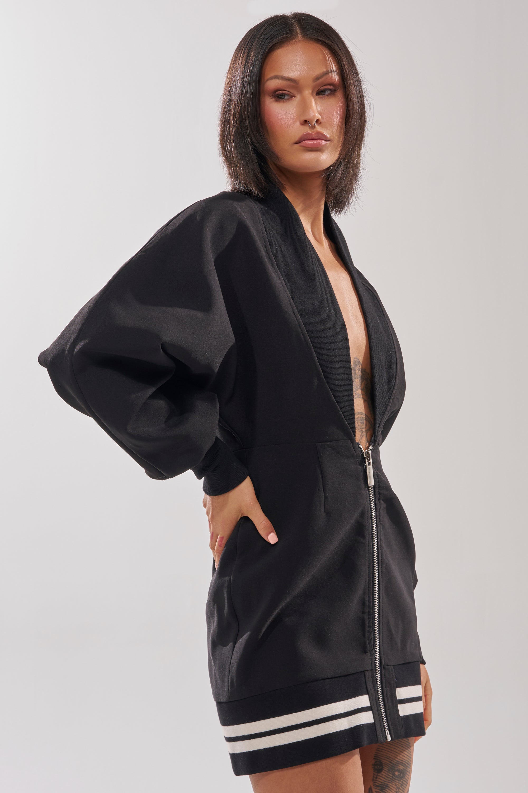Wearing the FAST LANE MINI DRESS—a black zip-up mini dress with wide sleeves and white striped trim—a woman with straight shoulder-length hair stands confidently, one hand on her hip, against a plain background.