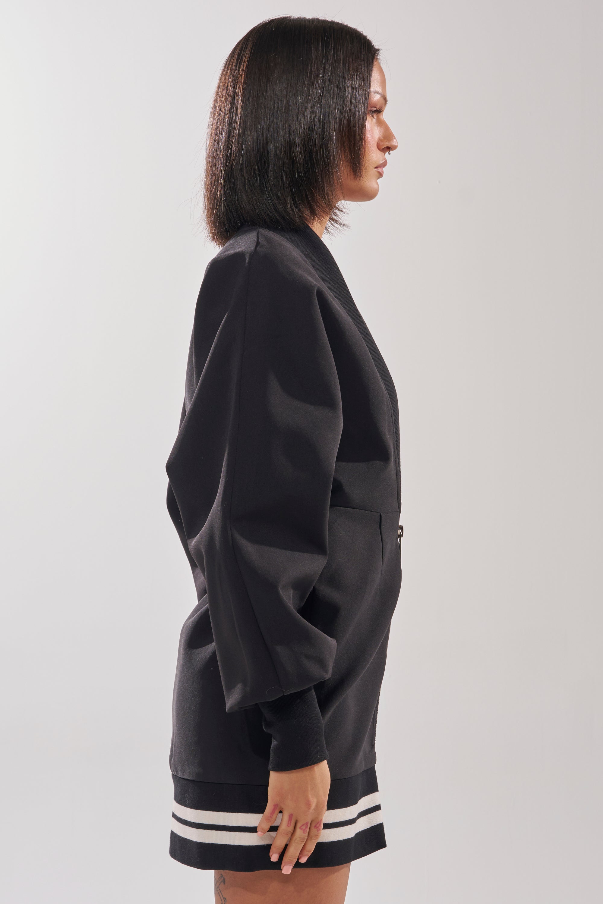 A woman with straight shoulder-length hair stands in profile against a plain light background, wearing the FAST LANE MINI DRESS—a sporty-chic black blazer-style mini dress with varsity-inspired white stripes near the hem.