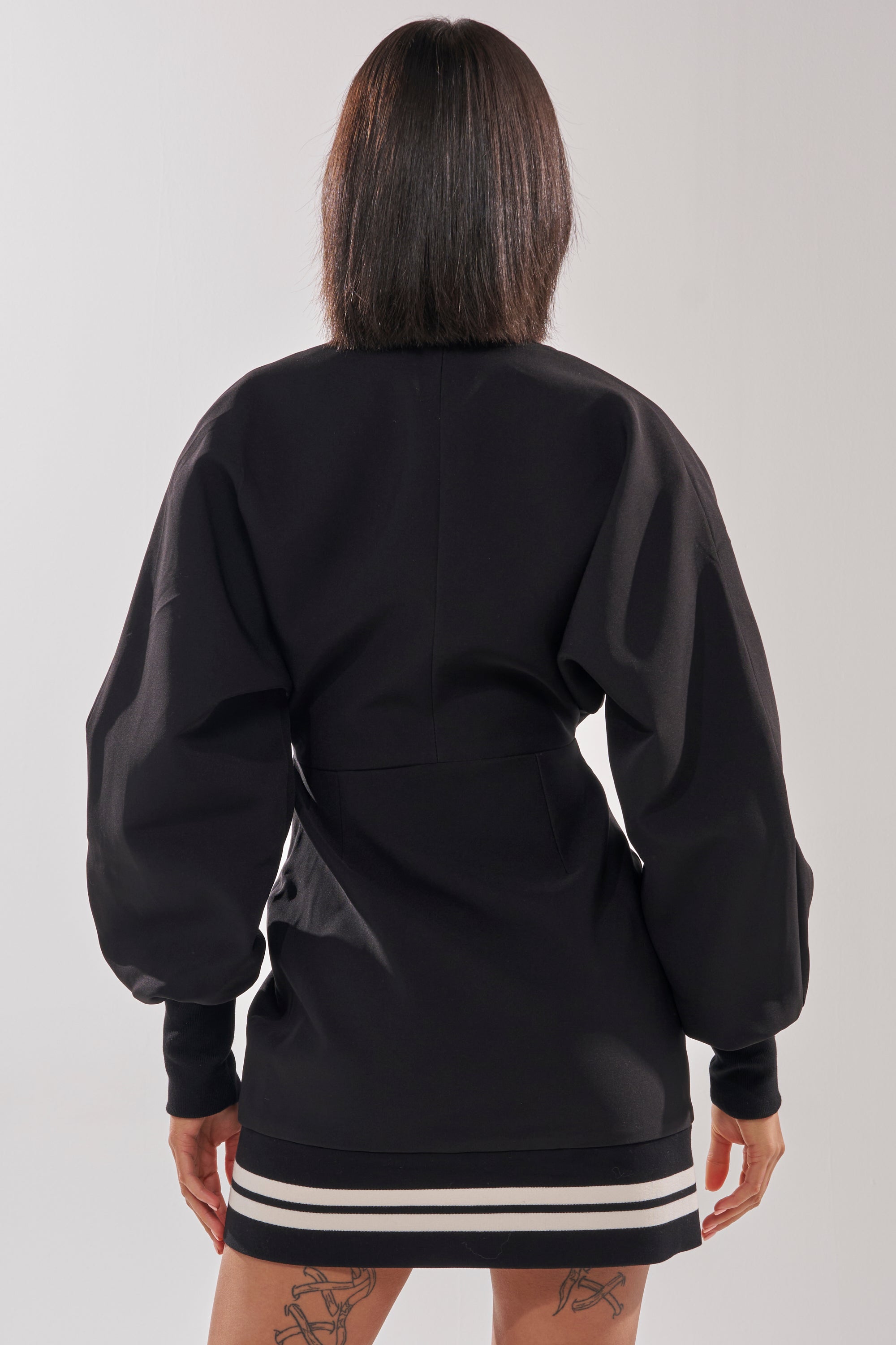 A person with shoulder-length dark hair is seen from the back wearing the FAST LANE MINI DRESS, a black varsity-inspired style with white stripes at the hem. Their hands rest by their sides, showing tattoo designs on both thighs.