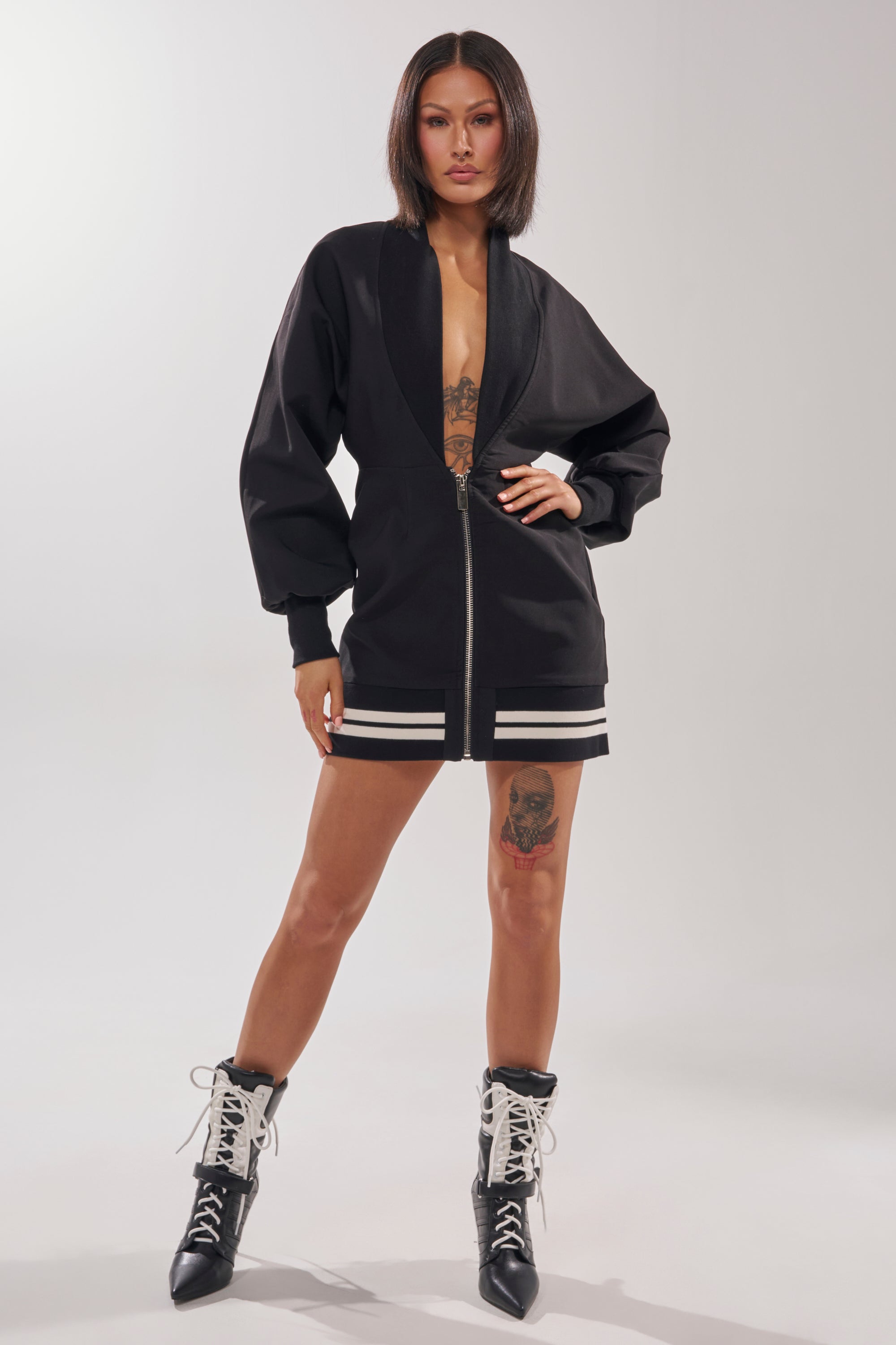 A woman with a bob haircut confidently wears the FAST LANE MINI DRESS, a black zip-up mini dress with white striped hem, paired with black lace-up ankle boots and showing tattoos on her chest and thigh.