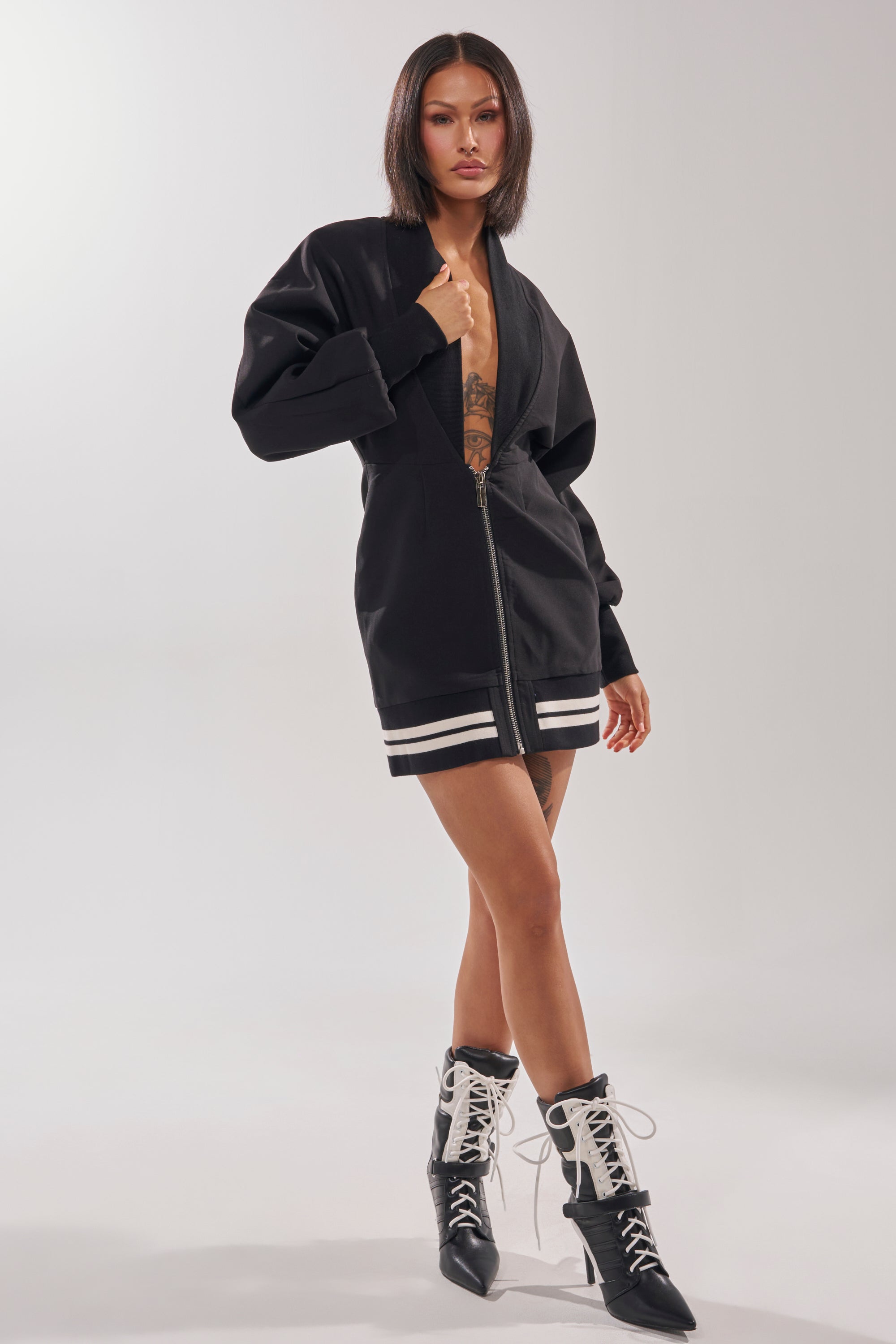A woman with straight dark hair models the FAST LANE MINI DRESS, a black varsity-inspired zip-up mini with white stripes on the hem and cuffs, paired with black lace-up ankle boots against a light, neutral background.