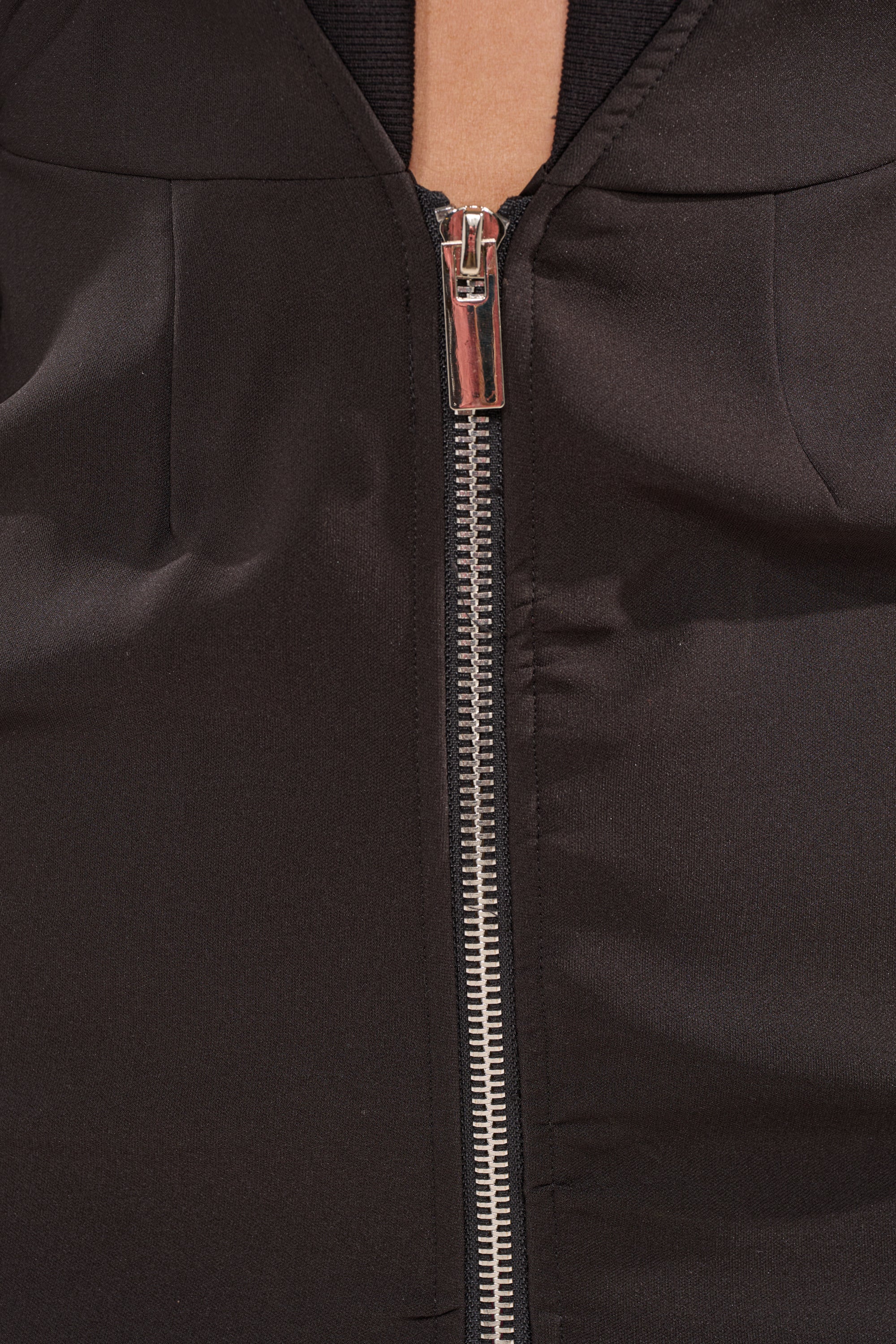 Close-up of the metallic zipper on the FAST LANE MINI DRESS, shown partially unzipped to reveal a hint of skin. The black fabric has a smooth, slightly shiny finish. 8