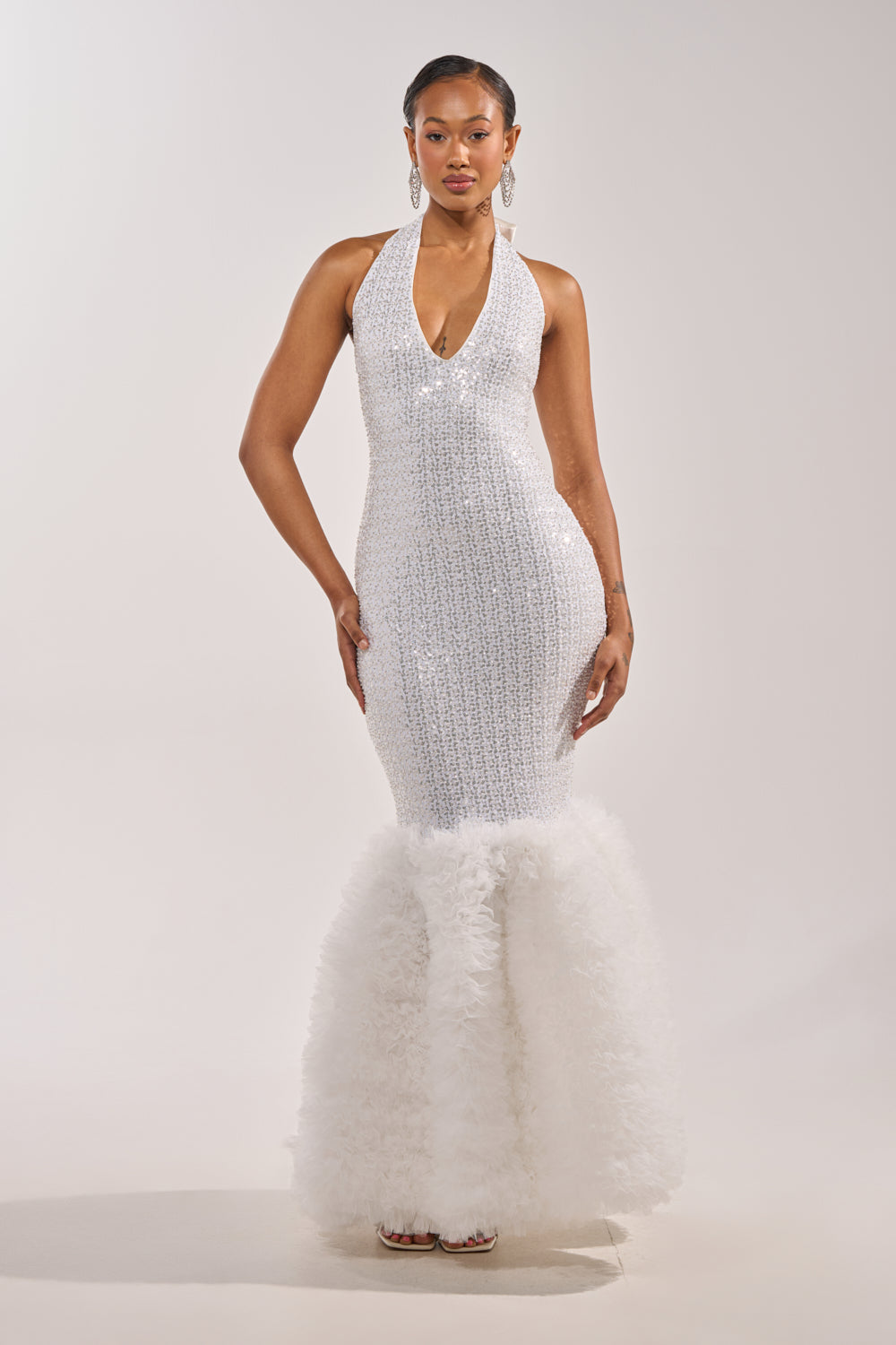 A woman models the SHOWTIME SEQUIN MAXI DRESS—a sleeveless, white mermaid gown with a deep V-neckline, sequin details, and a dramatic ruffled hem—standing against a plain light background. 0