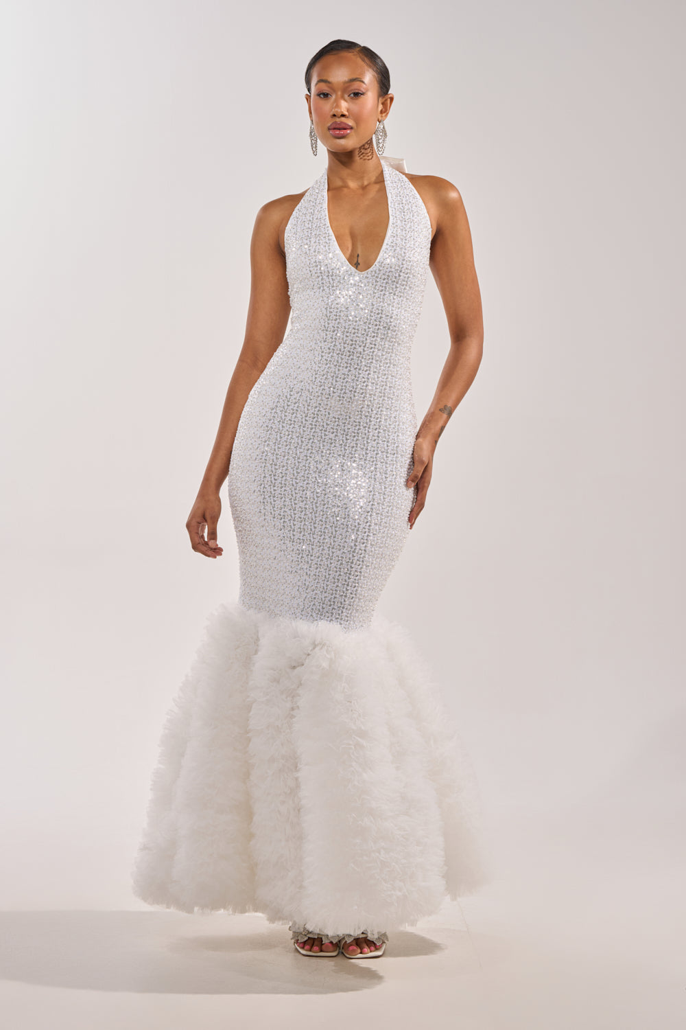 A woman poses confidently against a plain, light background wearing the SHOWTIME SEQUIN MAXI DRESS, a sleeveless white mermaid gown featuring a halter neckline and a voluminous ruffled skirt. 1