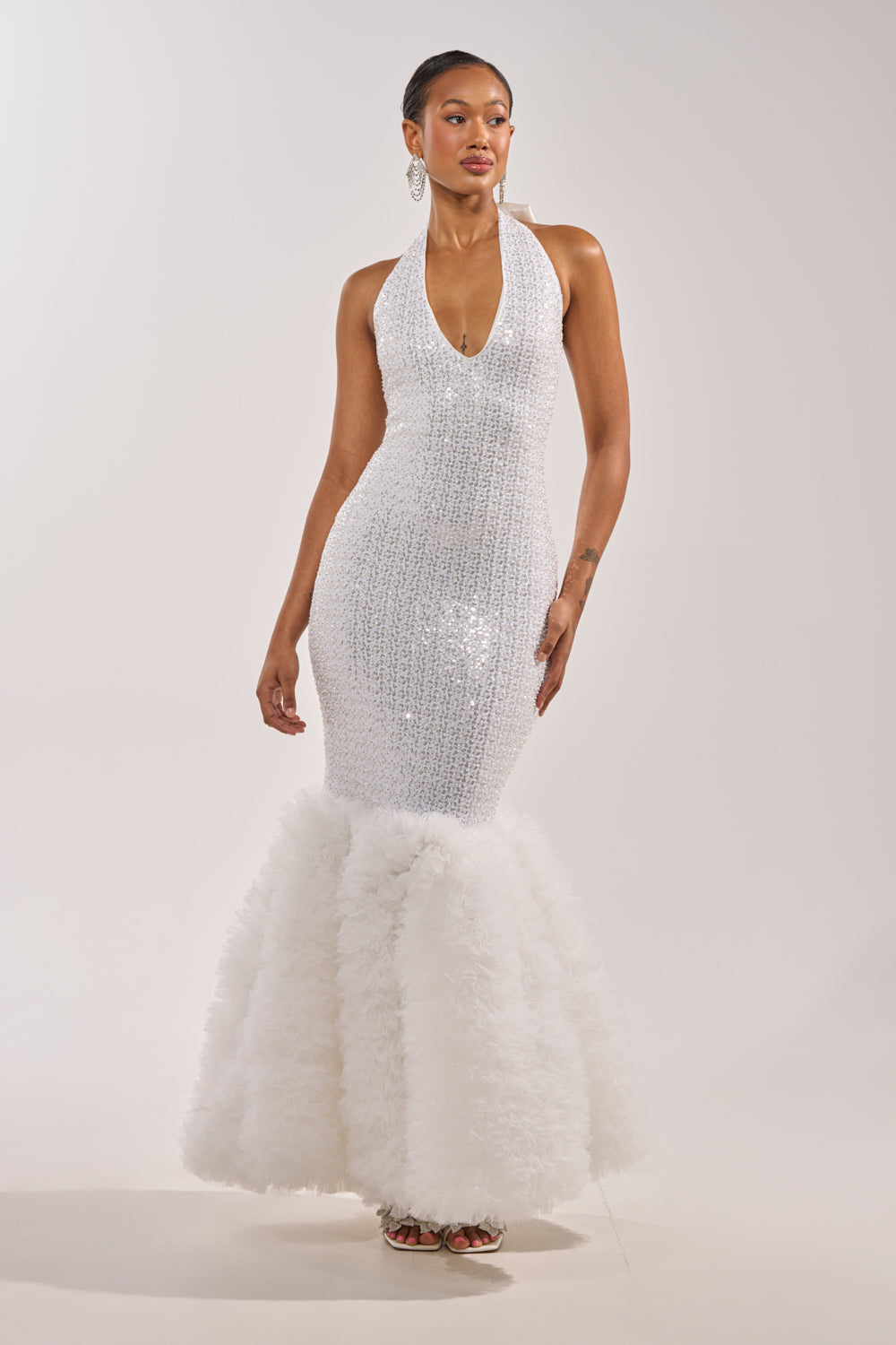 A woman stands against a plain light background in the SHOWTIME SEQUIN MAXI DRESS, a white mermaid gown with a deep V-neckline and ruffled hem. She wears hoop earrings and has sleek, styled-back hair. 2