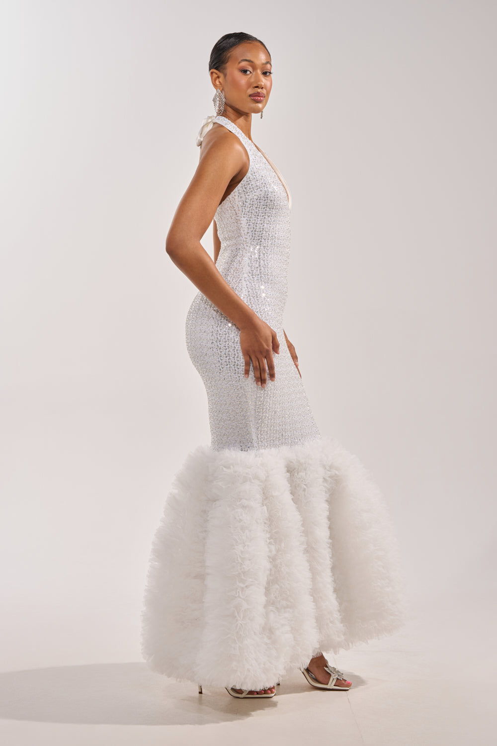 A woman poses in the SHOWTIME SEQUIN MAXI DRESS, a fitted white sequin mermaid gown with a dramatic ruffled hem. She accessorizes with earrings and white heels, standing against a plain light background. 3