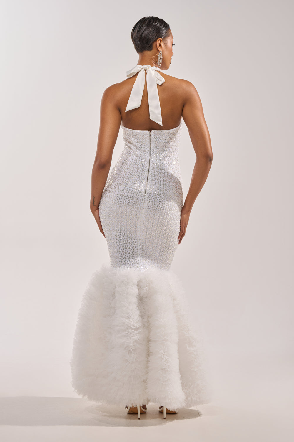 A woman faces away in the SHOWTIME SEQUIN MAXI DRESS, featuring a white sequin mermaid silhouette, halter neckline, sparkling bodice, and a dramatic feathered skirt. She wears her hair sleek and short with bold statement earrings. 4