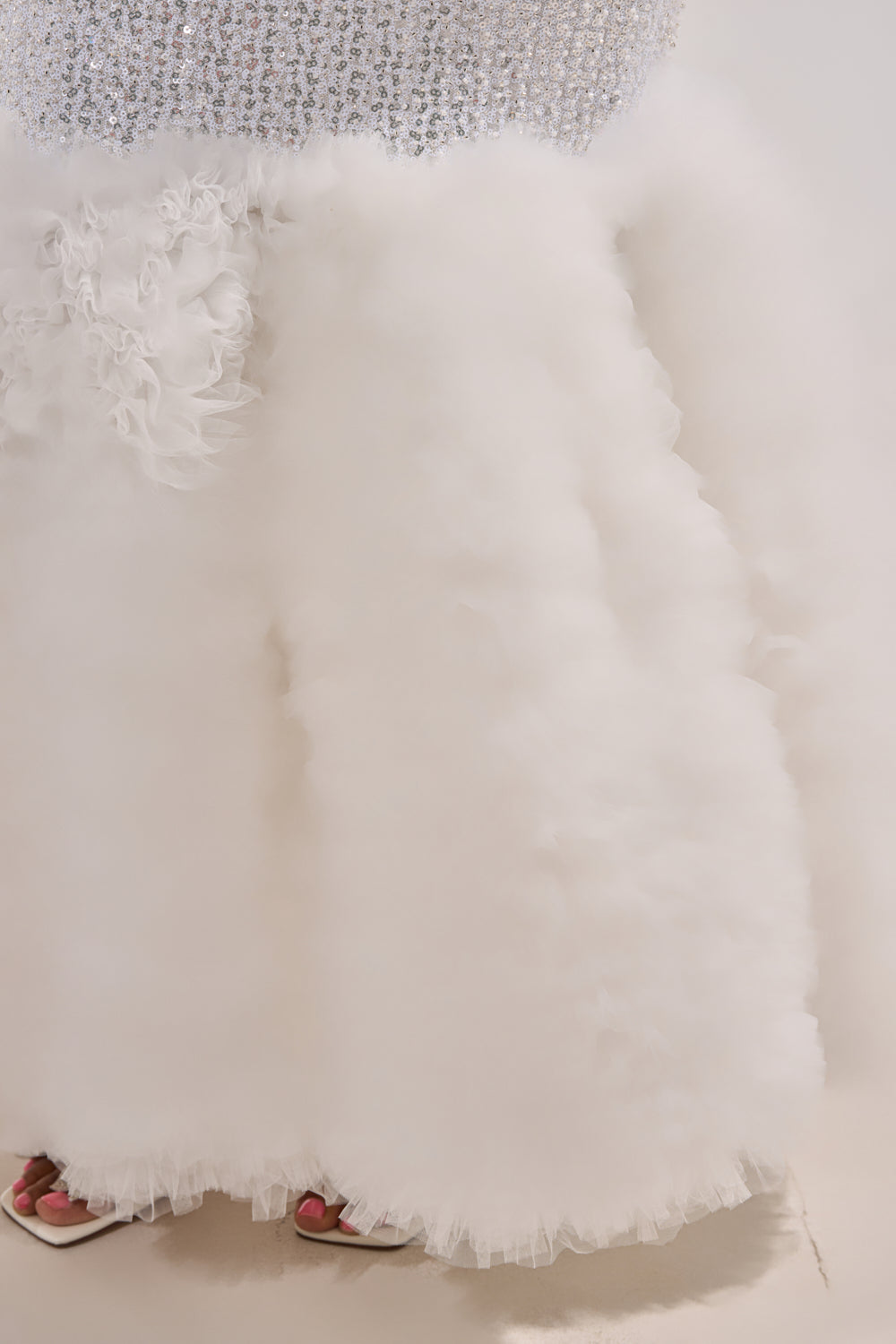 Close-up of the SHOWTIME SEQUIN MAXI DRESS with a silver sequined bodice and voluminous ruffled tulle skirt, paired with white heeled sandals and pink nail polish peeking from under the hem. 6