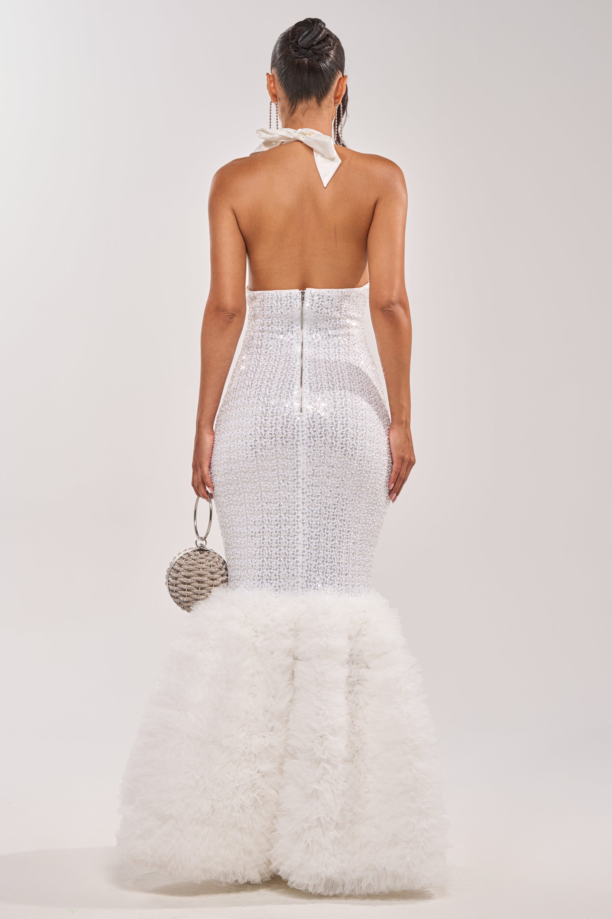 A woman faces away in the SHOWTIME SEQUIN MAXI DRESS, a dazzling white halter mermaid gown with a feathered hem, ideal for a red carpet look. She holds a small round purse and wears her hair in a sleek ponytail. 6