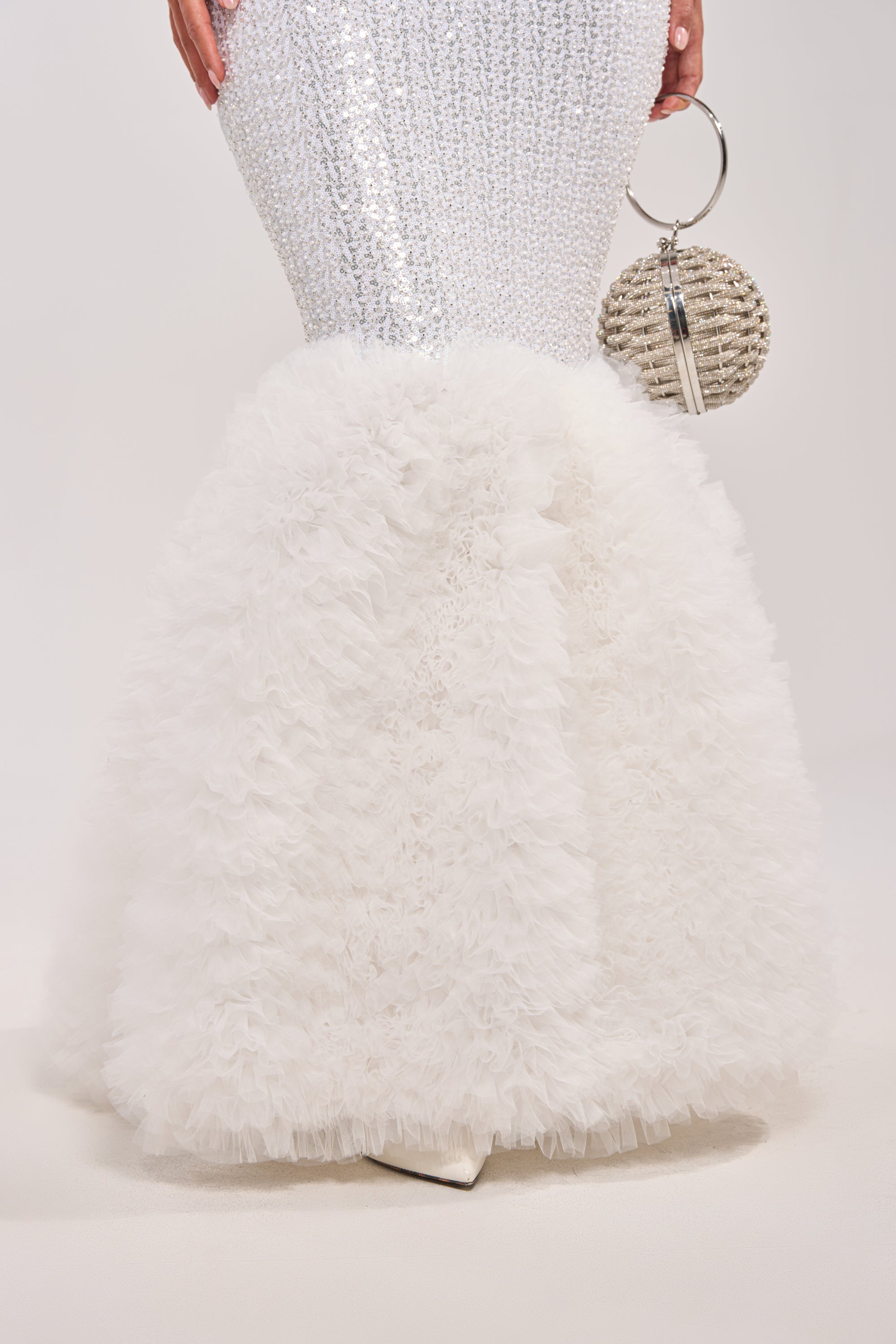 A close-up of the lower half of the SHOWTIME SEQUIN MAXI DRESS, a fitted white sequin mermaid gown with a ruffled tulle skirt. The person holds a textured round silver clutch featuring a large ring handle. 7