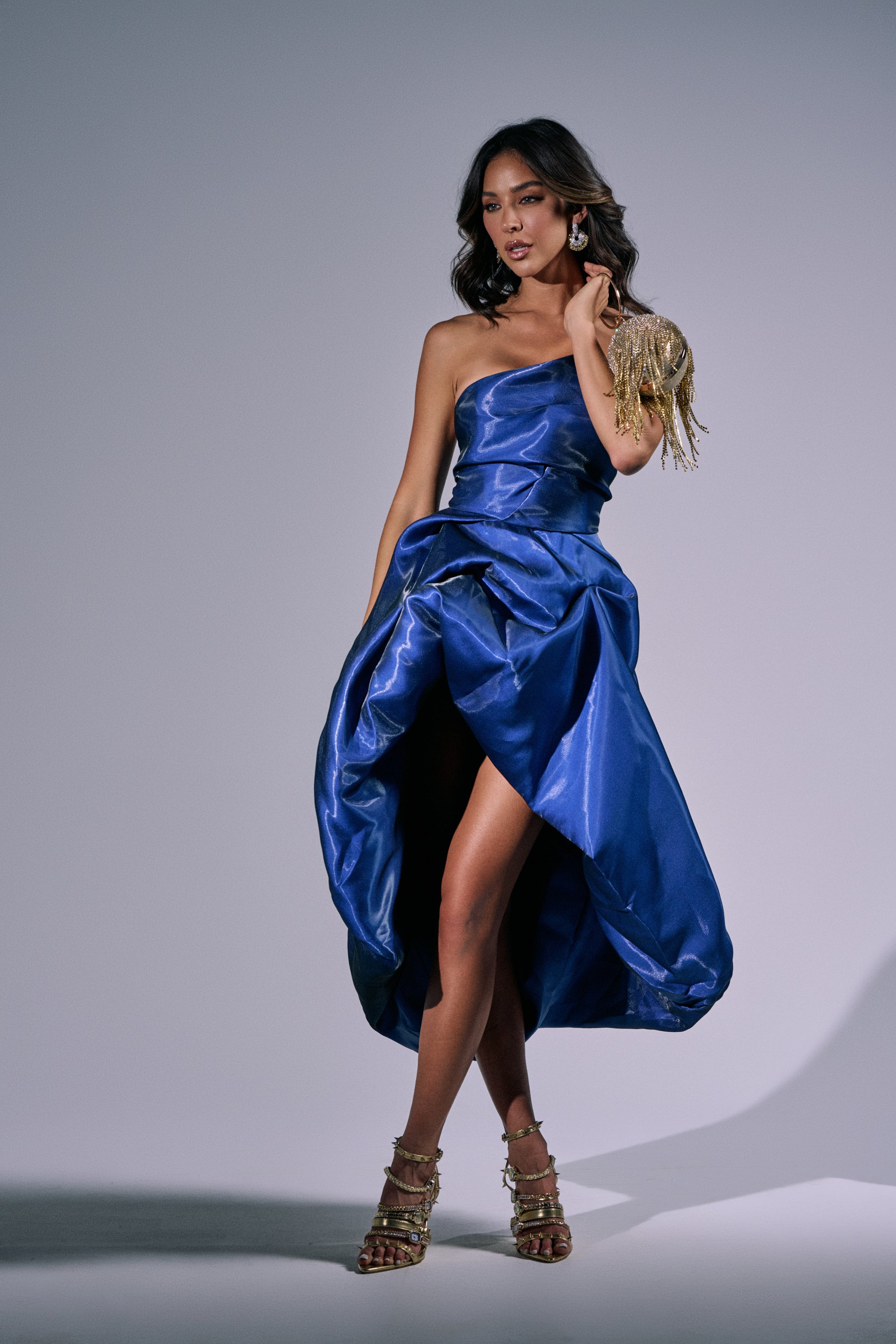 A woman models the HEAD OVER HEELS HIGH-LOW DRESS IN BLUE, featuring a couture-inspired design with a high slit and gold accents, paired with strappy gold heels and statement earrings against a plain gray background. 1