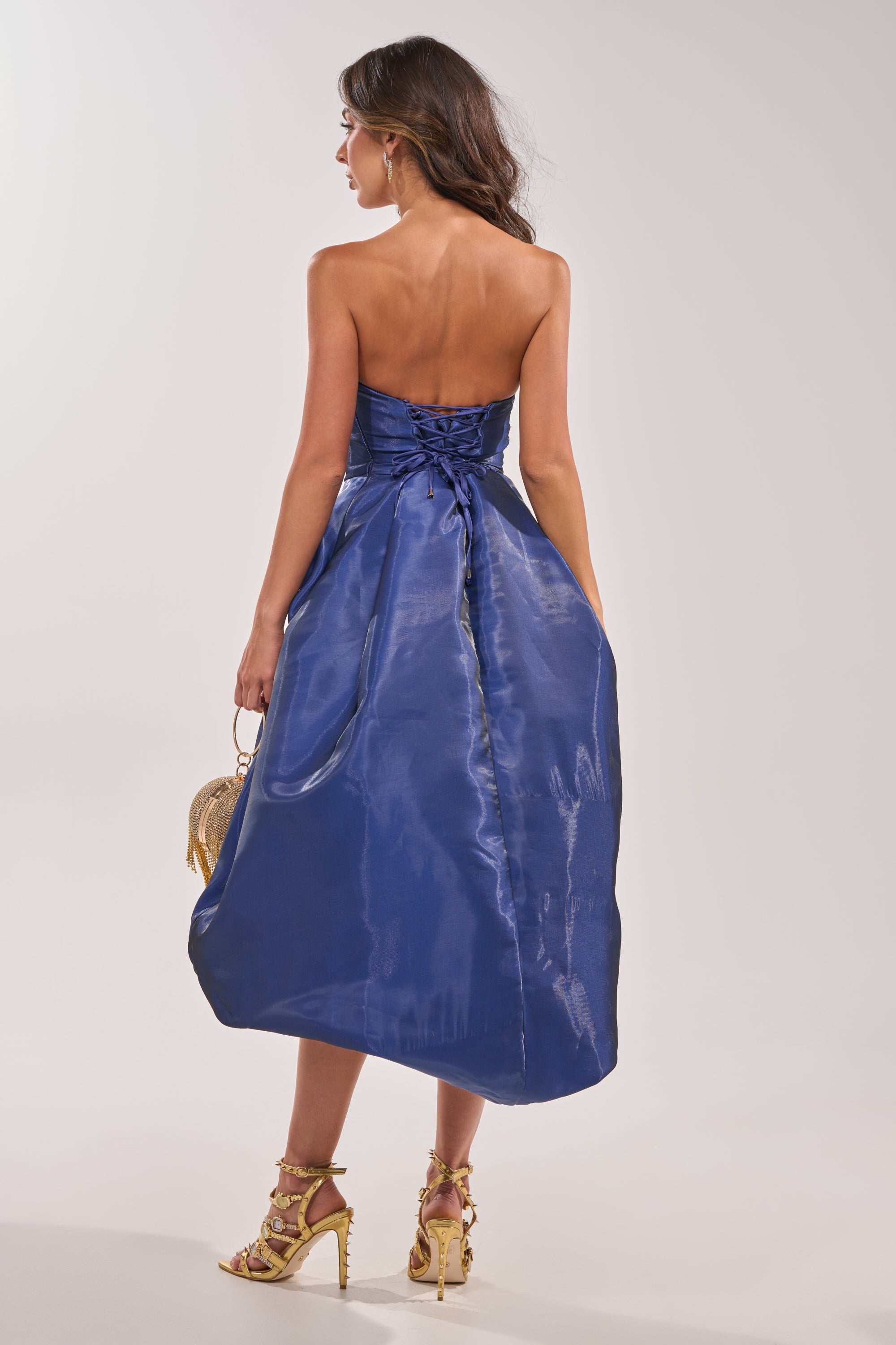 A woman faces away in the HEAD OVER HEELS HIGH-LOW DRESS IN BLUE, featuring a fitted bodice and flared skirt. She carries a small gold purse, wears strappy gold heels, and her loose waves complete the elegant evening look. 6