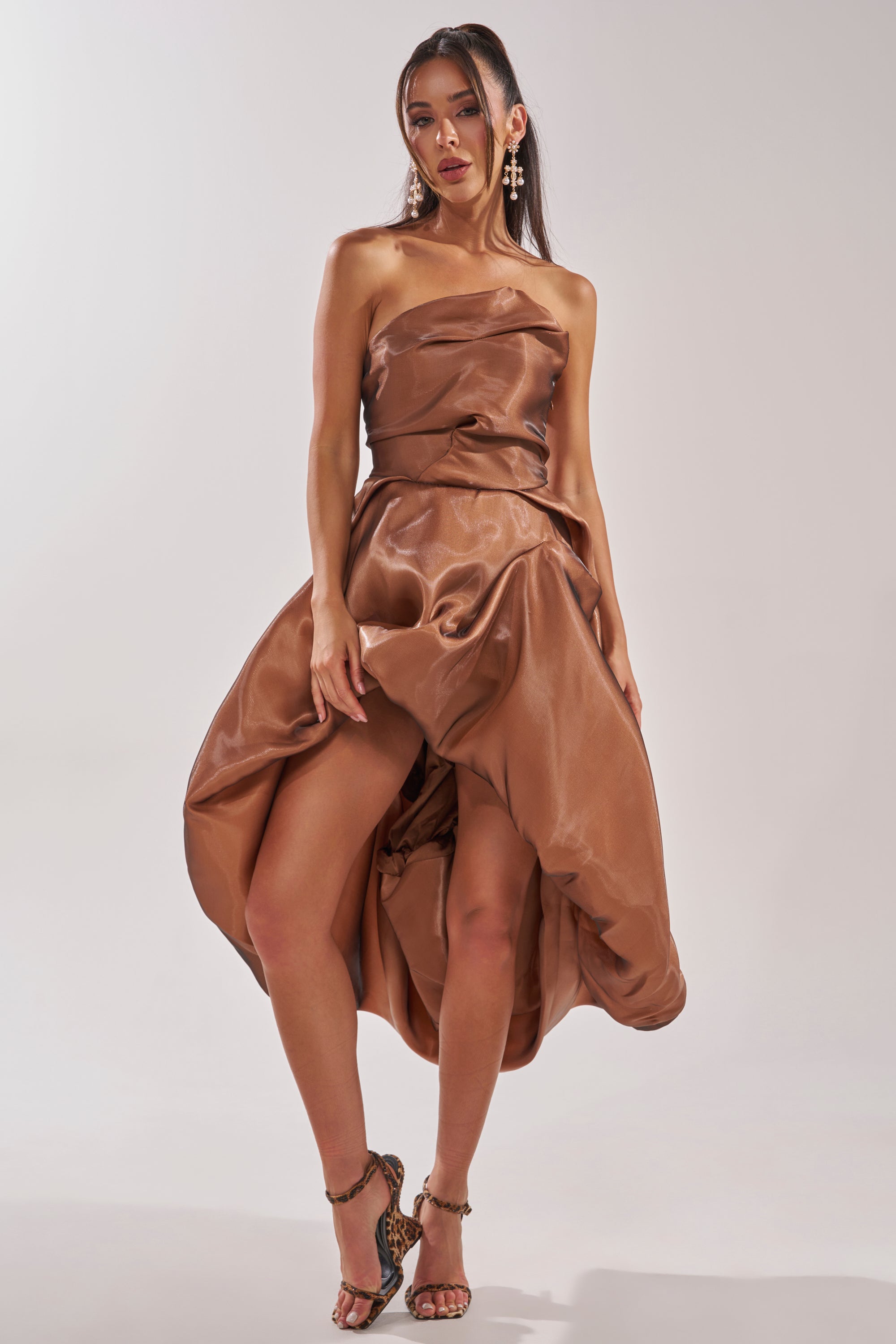 A woman poses confidently in the HEAD OVER HEELS HIGH-LOW DRESS, a strapless tulle cocktail dress in shiny brown-burgundy with a voluminous skirt. She accessorizes with bold earrings and strappy heels against a light background. 1