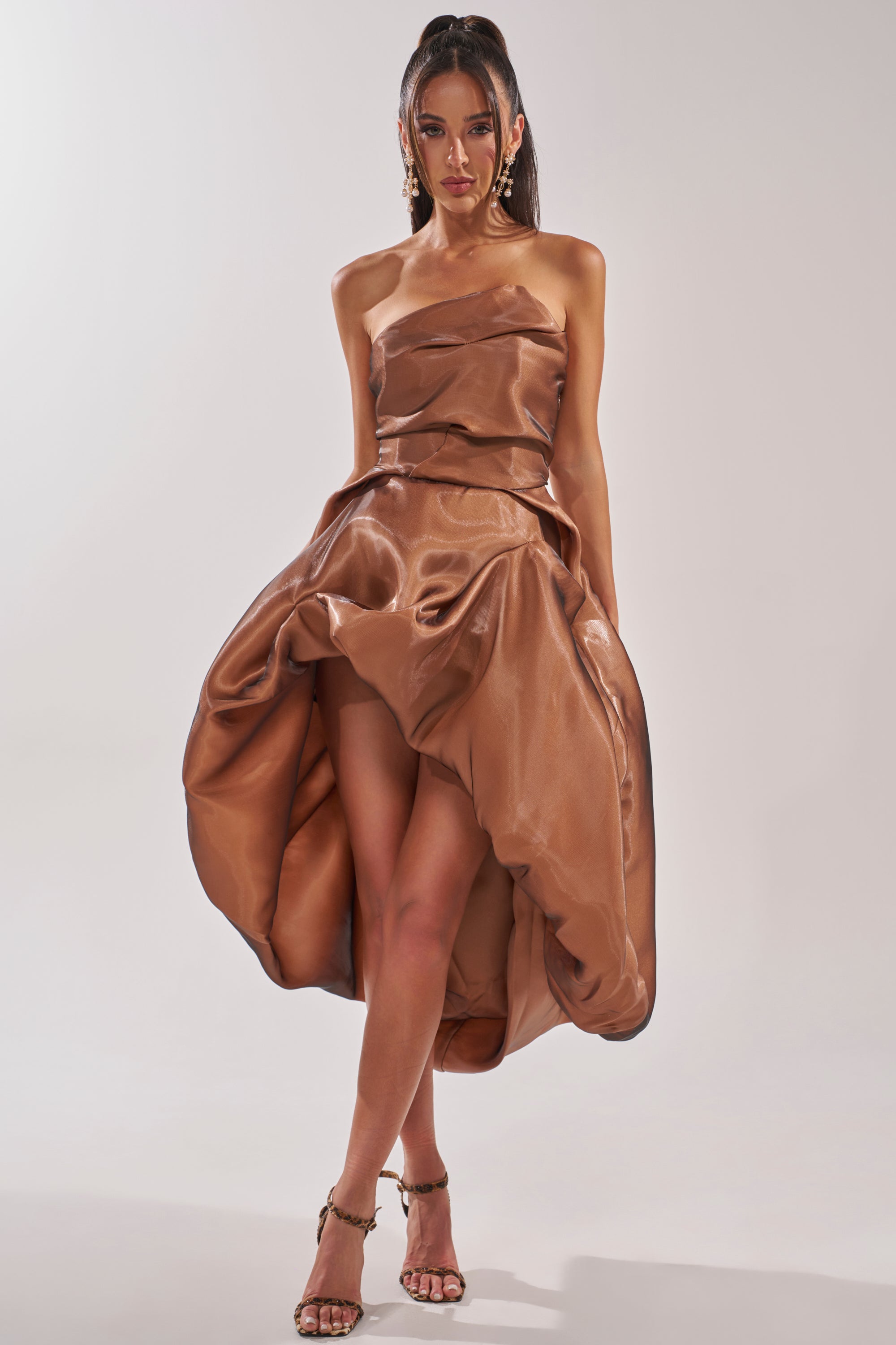A woman models the HEAD OVER HEELS HIGH-LOW DRESS—a strapless tulle cocktail dress with a shiny bronze high-low skirt. She pairs it with strappy heels, drop earrings, and a sleek high ponytail against a plain backdrop. 2