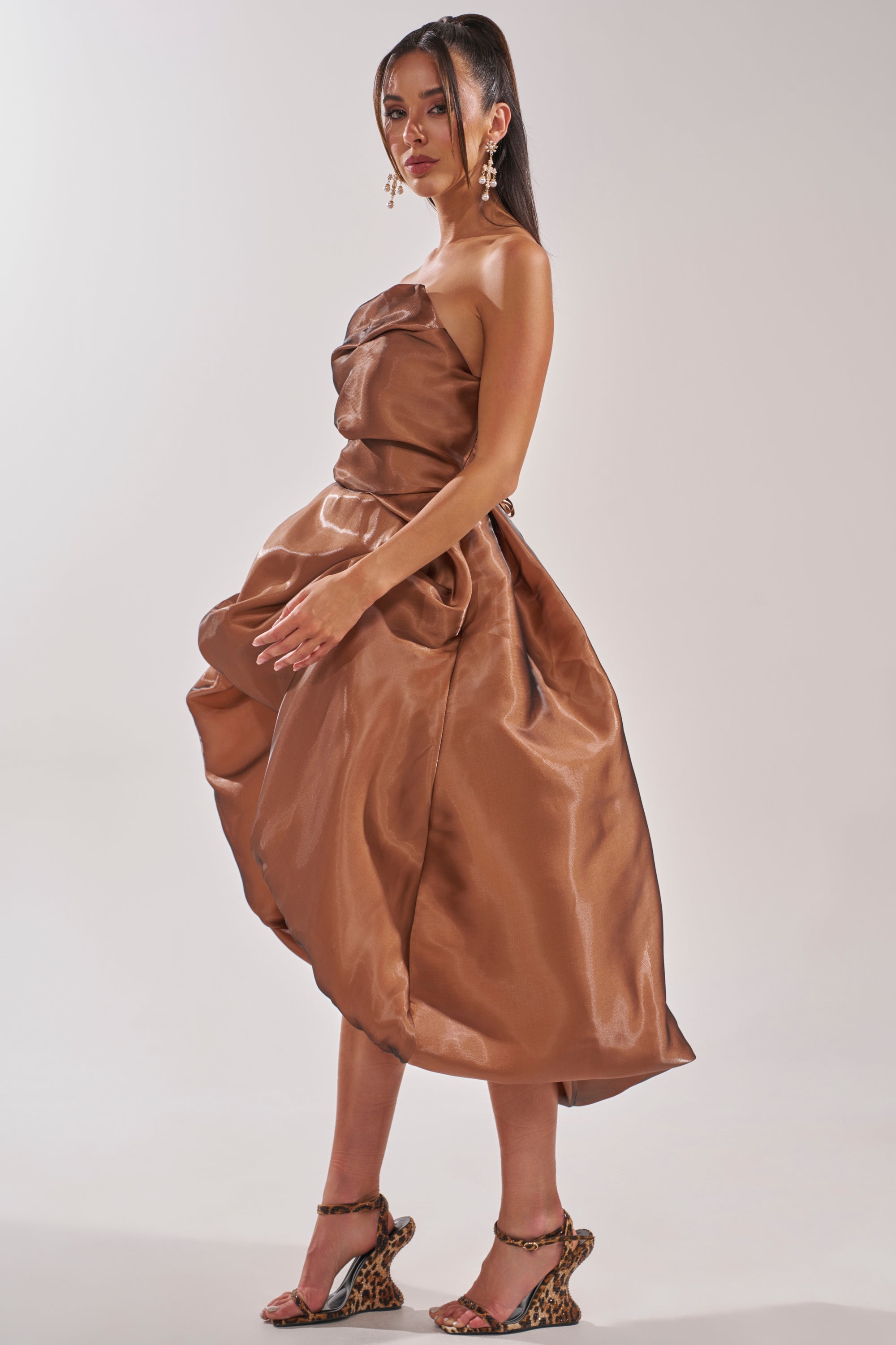 A woman poses in the HEAD OVER HEELS HIGH-LOW DRESS, a shiny, strapless formal gown with a voluminous skirt. She completes the look with statement earrings, high ponytails, and leopard-print heeled sandals against a plain background. 3