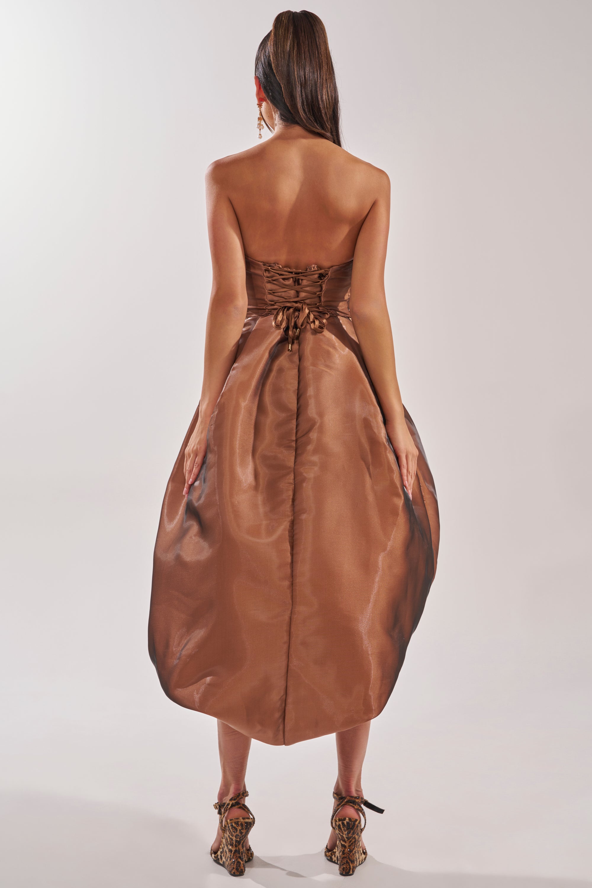 A woman faces away, wearing the HEAD OVER HEELS HIGH-LOW DRESS with a strapless, corset-style lace-up back and voluminous skirt. Her long ponytail pairs with patterned high-heeled shoes. 5