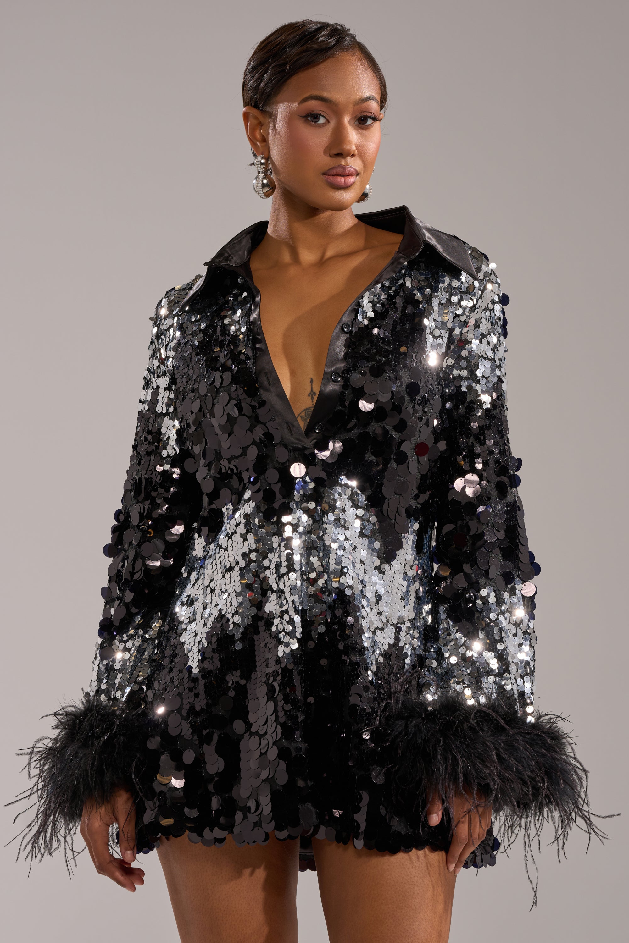 A woman poses confidently in the SEQUINS AFTER DARK MINI DRESS—a black sequin mini dress with long sleeves, feathered cuffs, and a collar—perfect for elegant holiday parties. She wears earrings, her short hair styled, against a plain background.