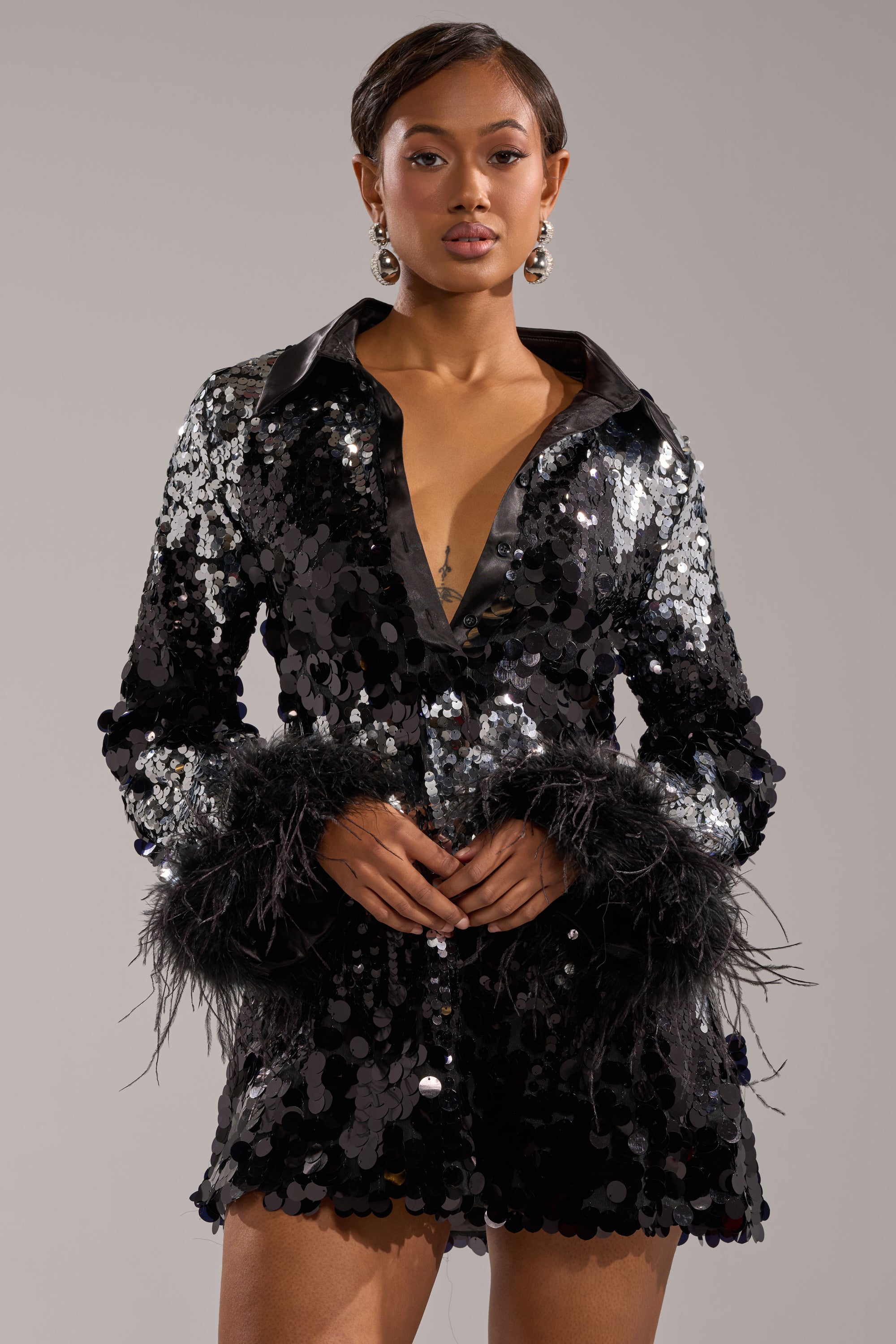 A woman poses confidently in the SEQUINS AFTER DARK MINI DRESS—featuring black feathers, sparkling sequins, a deep V-neckline, and statement earrings—against a grey background. This dress is a stunning choice for any holiday party.