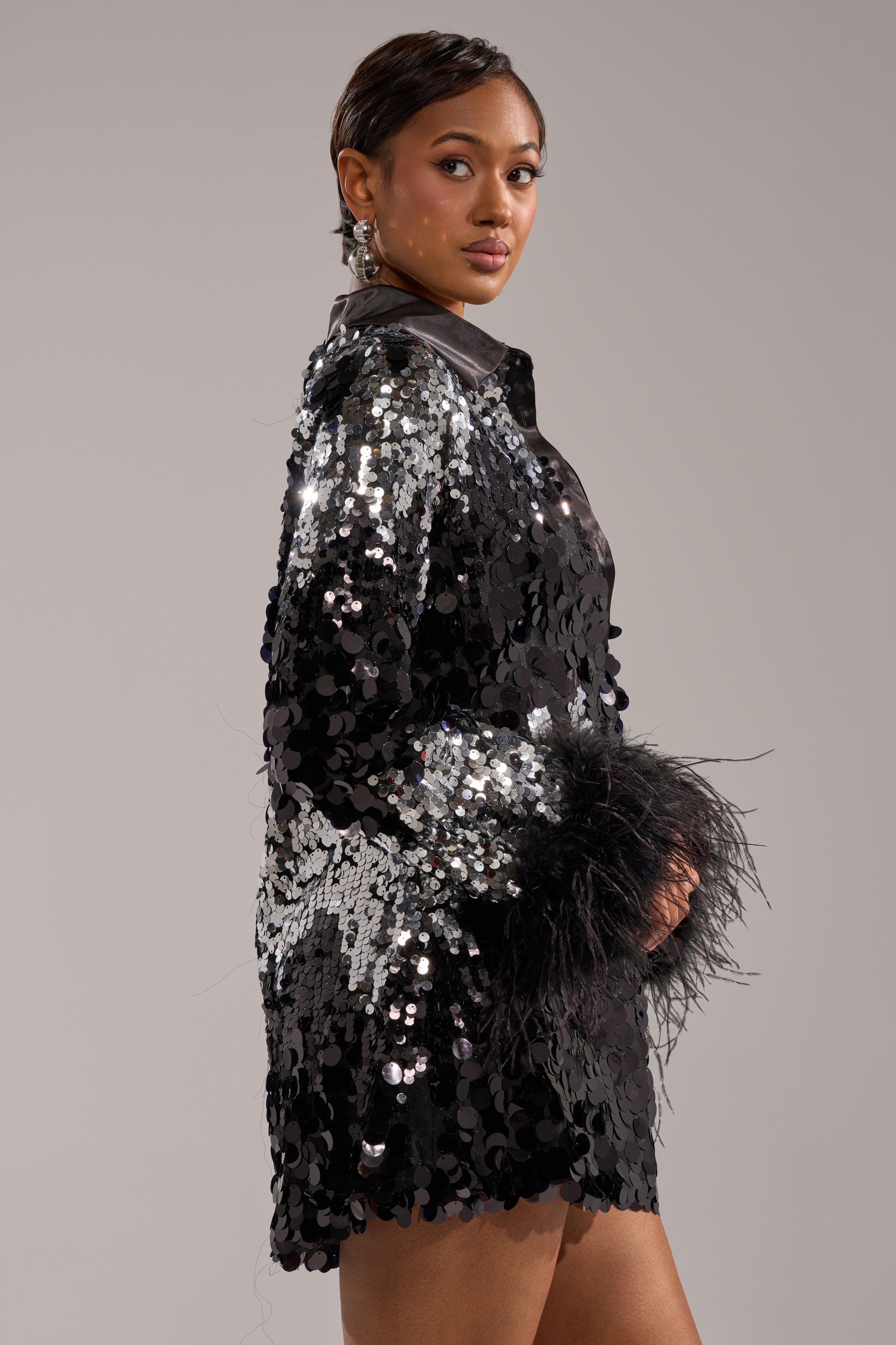 A woman poses confidently in the SEQUINS AFTER DARK MINI DRESS, looking over her shoulder. The plain, neutral background highlights her and the sparkling black sequin mini dress with silver accents.