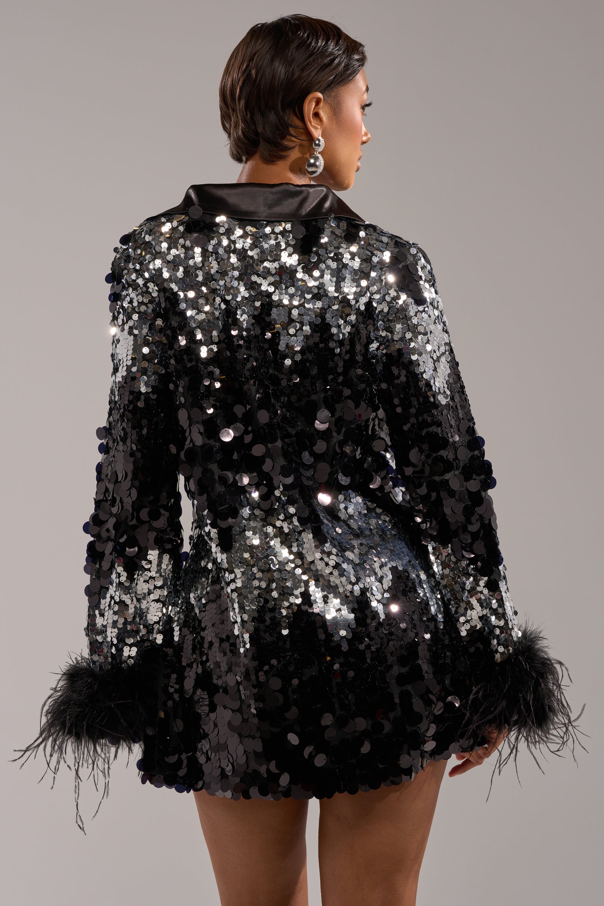 A person faces away, wearing the SEQUINS AFTER DARK MINI DRESS—a black sequin mini with reflective discs and feather cuffs. With short hair and large hoops, they pose against a light gray background, ideal for holiday party style.