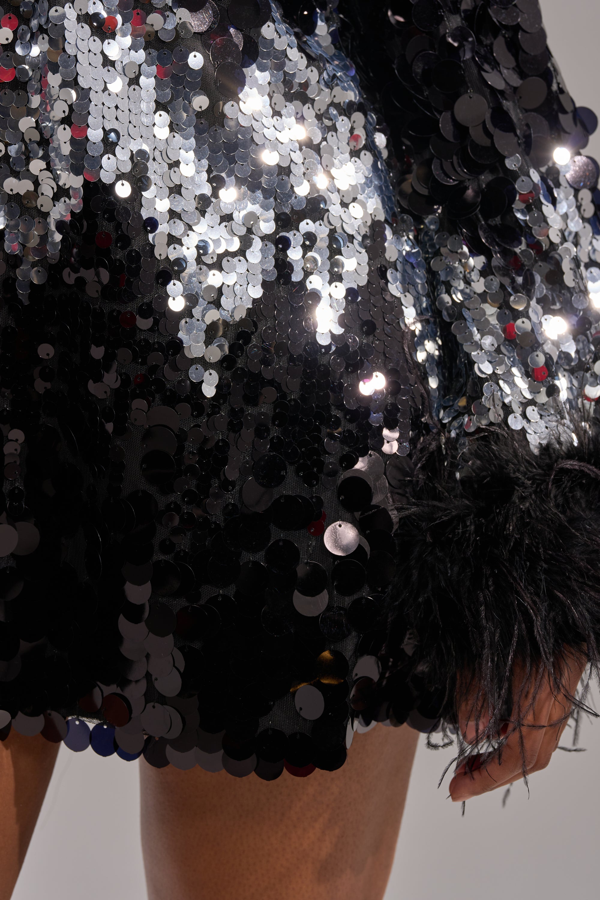 Close-up of a hand near the SEQUINS AFTER DARK MINI DRESS, featuring black feather cuffs and shiny silver sequins that reflect light for a sparkling effect.