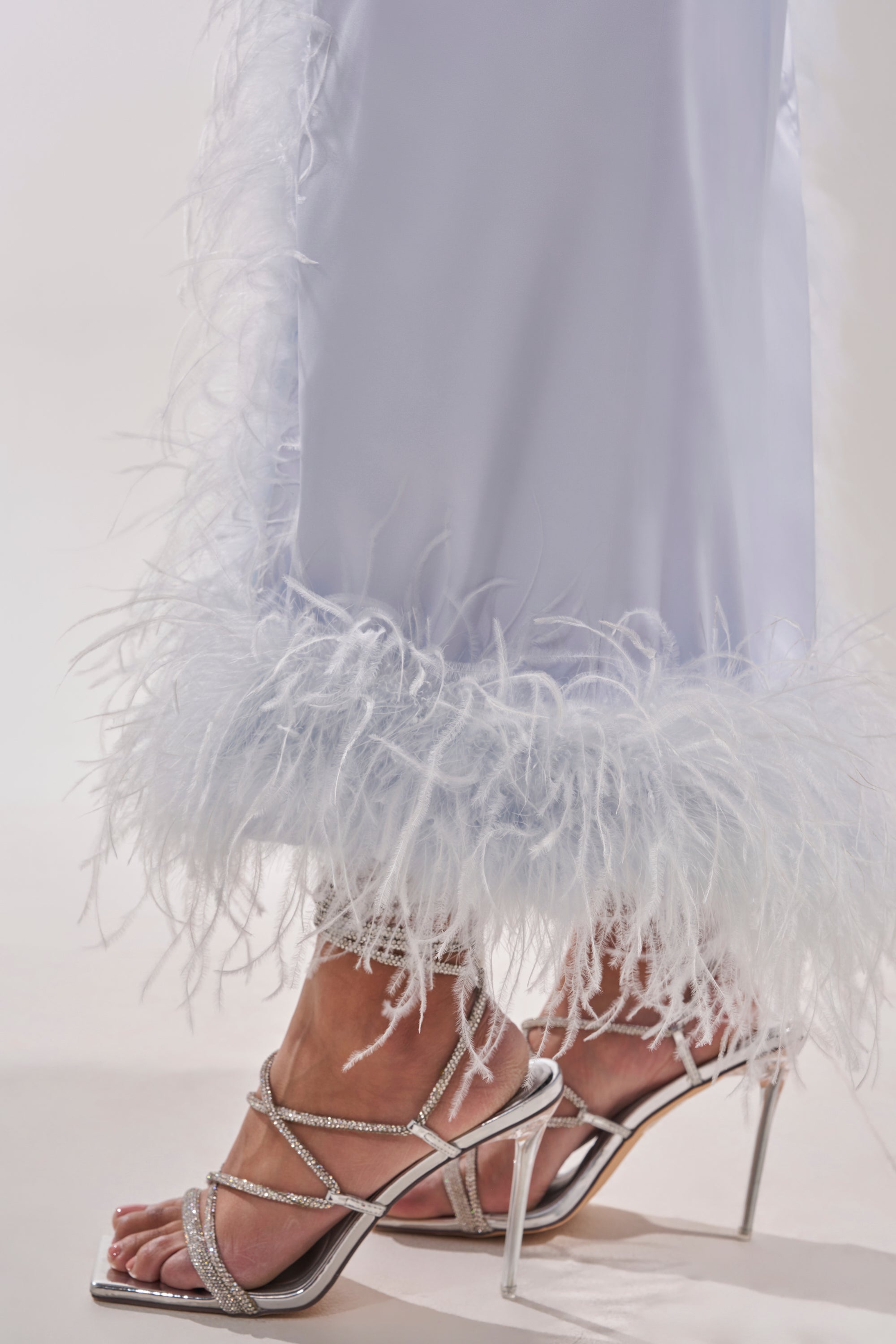 A close-up of a person wearing silver high-heeled sandals with rhinestone straps and the FROSTBITE MAXI DRESS—a pale lavender halter evening gown with fluffy feather trim at the hem. 10