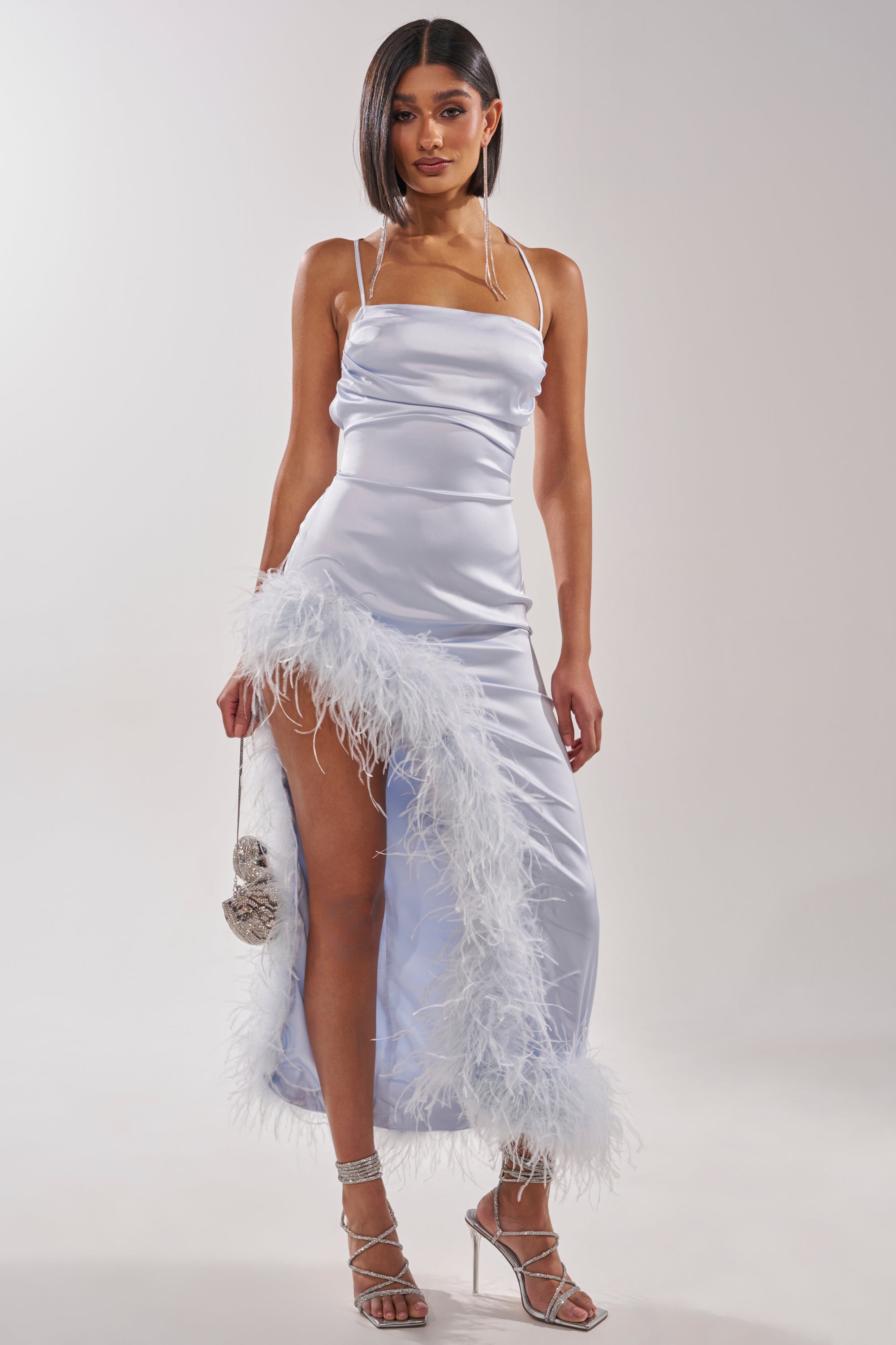 A woman models the FROSTBITE MAXI DRESS, a sleek white satin halter gown with feathery hem trim and thigh-high slit, styled with silver strappy heels and a sparkly clutch against a plain light background. 1