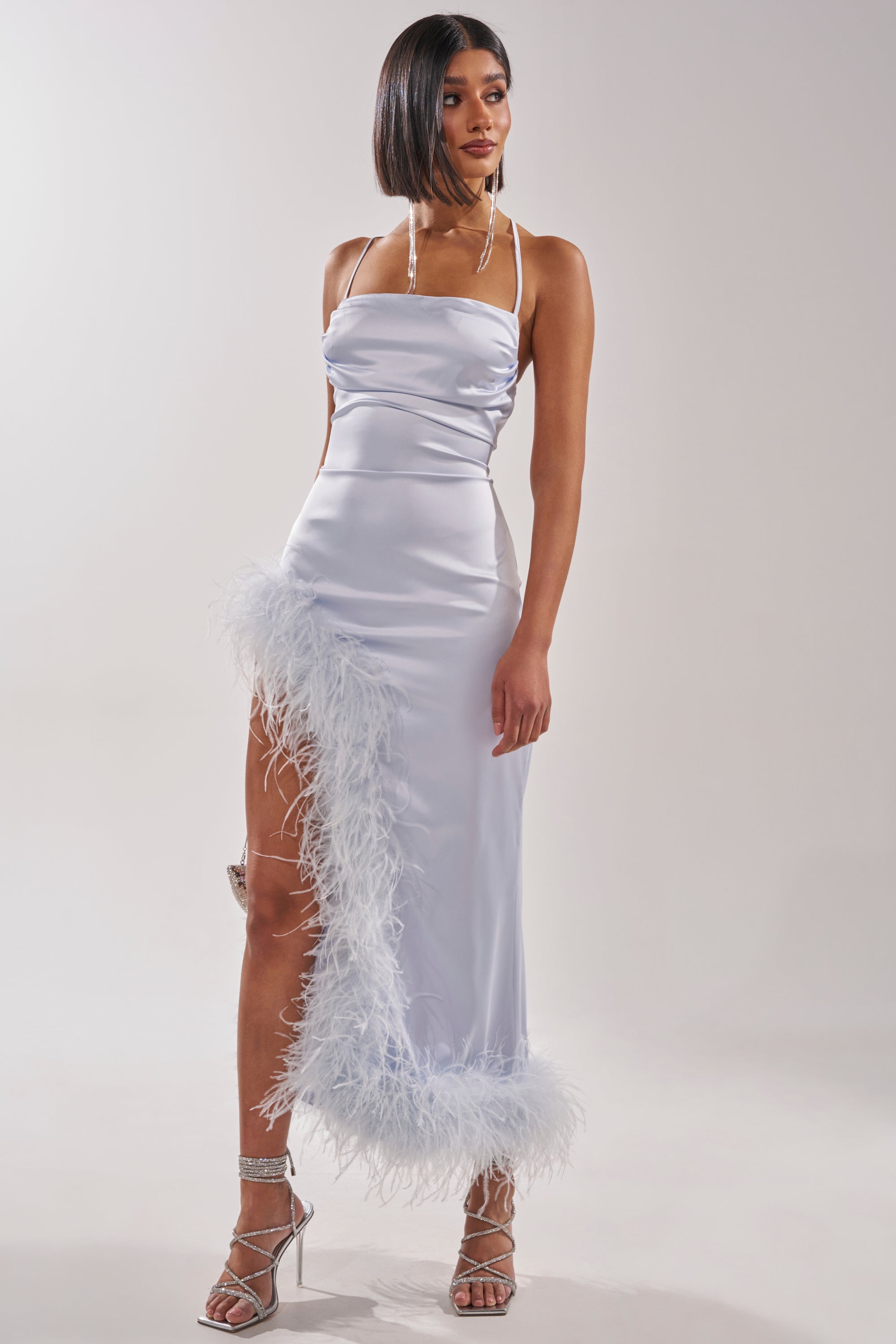 A woman stands against a plain background wearing the FROSTBITE MAXI DRESS, a light blue formal evening gown with feather-trimmed hem, thigh-high slit, and silver strappy heels. 2