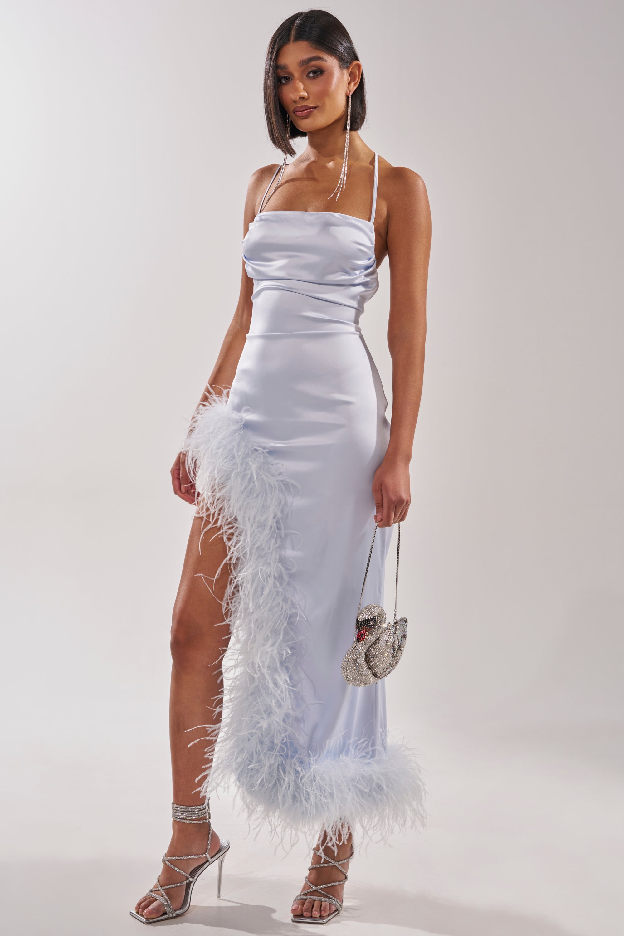 A woman wears the FROSTBITE MAXI DRESS, a light purple satin halter evening gown with white ostrich feather trim and a thigh-high slit, styled with strappy heels and a silver purse against a plain light background. 3