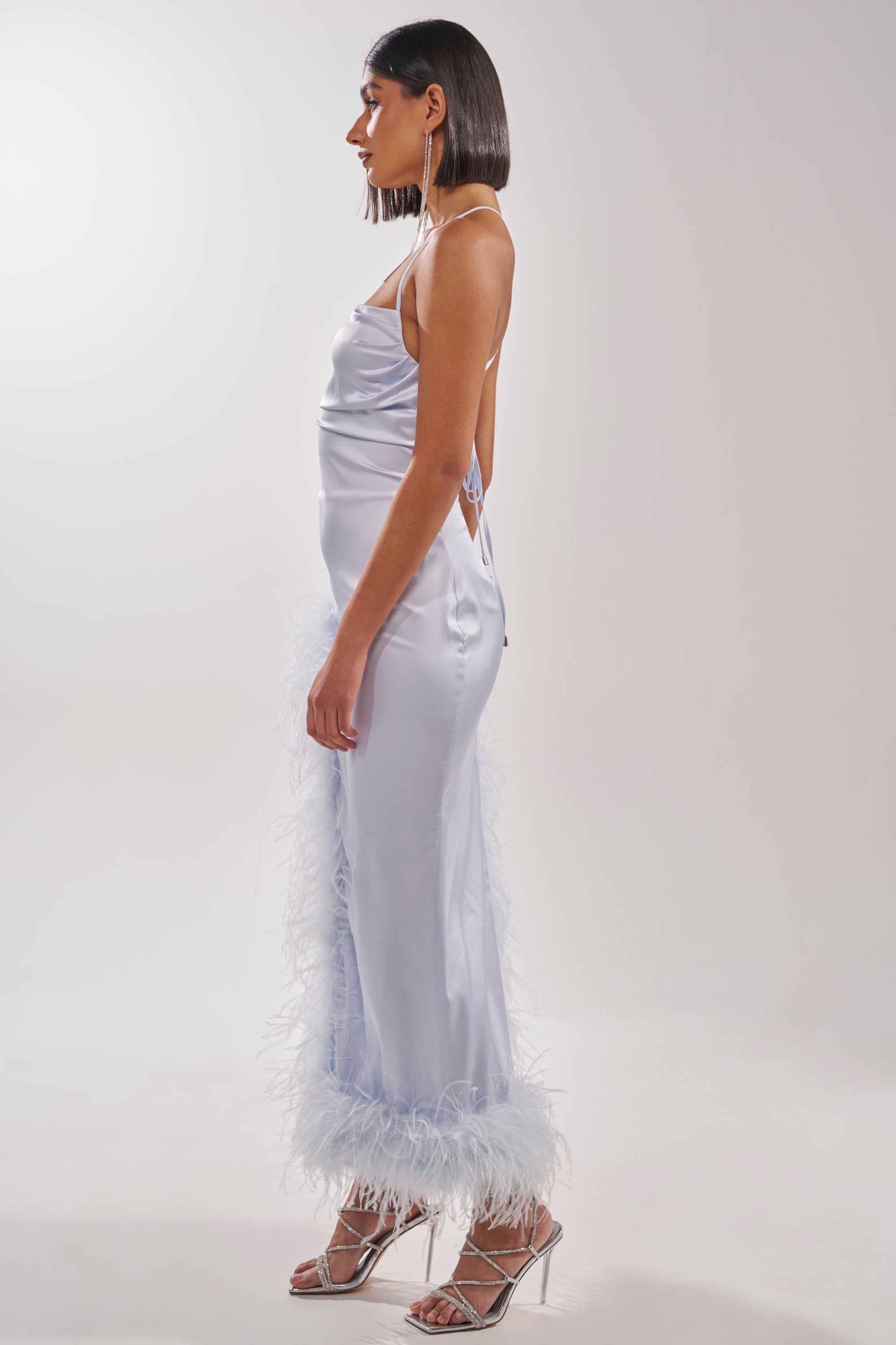 A woman in profile wears the FROSTBITE MAXI DRESS, a light blue evening gown with ostrich feather trim at the hem. She has straight, shoulder-length dark hair and silver high-heeled sandals, posing against a plain light background. 4