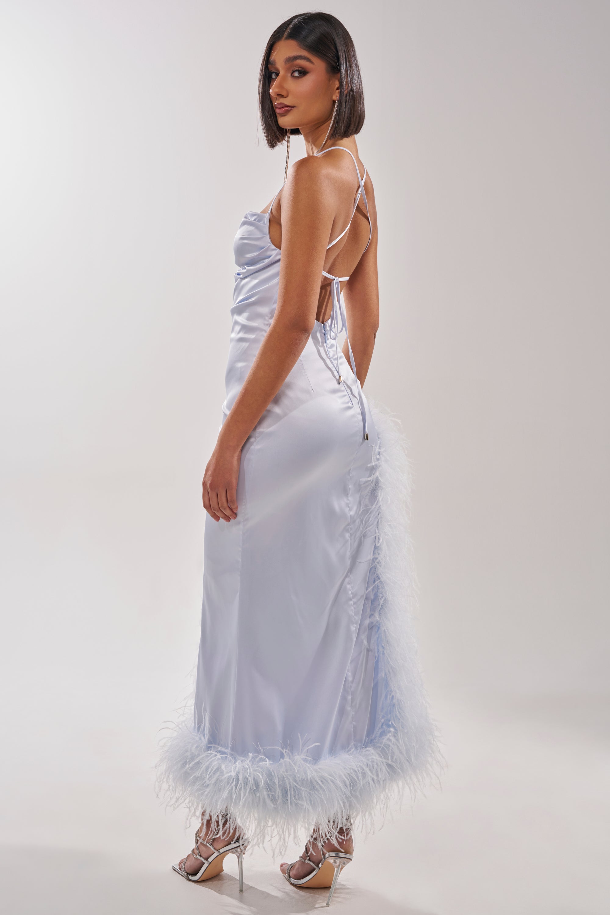 A woman in the FROSTBITE MAXI DRESS—a blue ostrich feather gown with thin straps and a feathered hem—stands in high heels, looking over her shoulder against a plain white background. 7