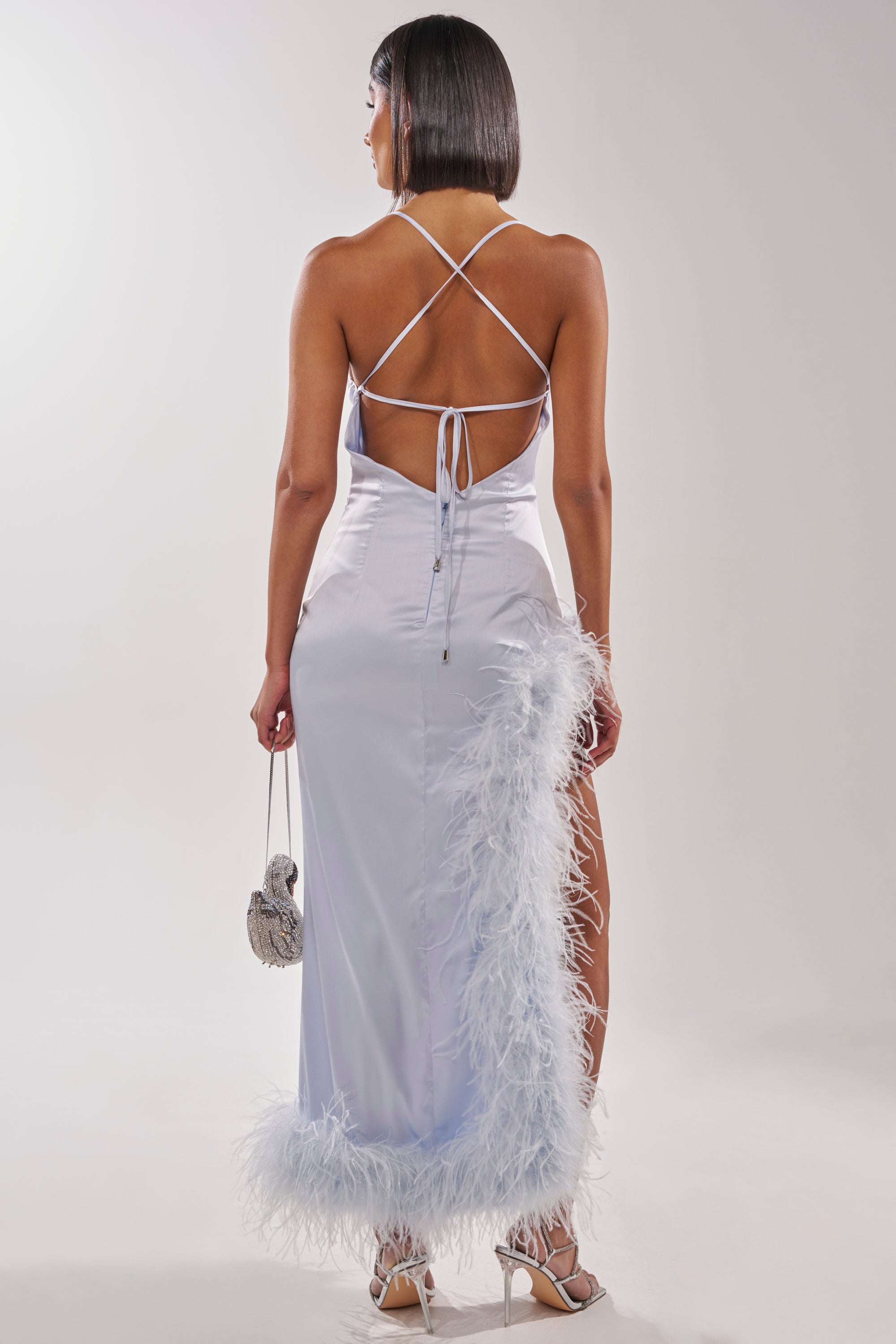 Wearing the FROSTBITE MAXI DRESS, a woman faces away, showcasing its silky light blue fabric and ostrich feather-trim. With crisscross straps, a silver purse, white heels, and a chic bob haircut, she’s styled perfectly for a formal gala. 8