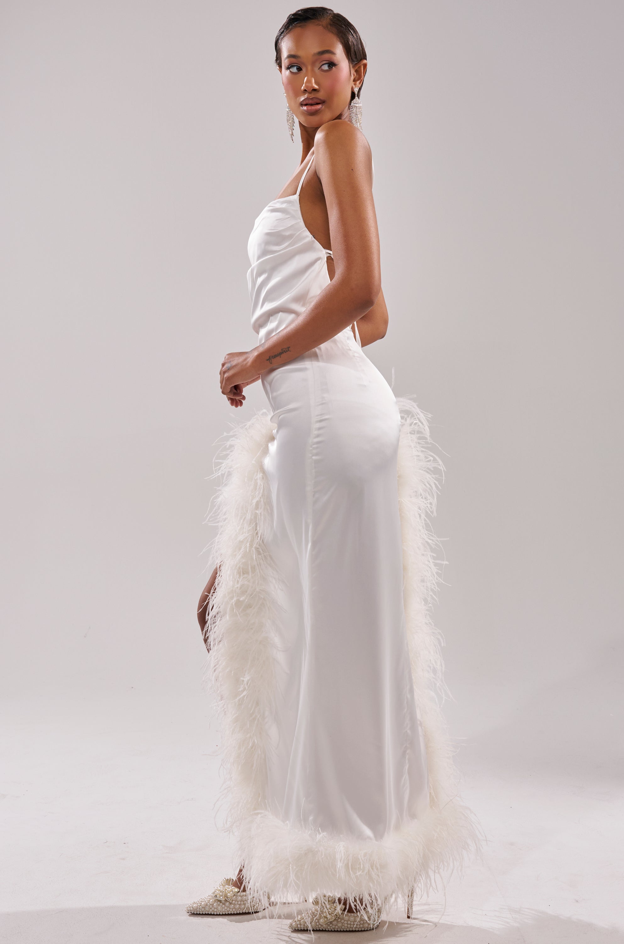 A woman stands in profile against a plain light background, wearing the LIGHT AS A FEATHER MAXI DRESS—crafted from white satin fabric with feather trim—paired with earrings and sparkly pointed shoes, gazing confidently at the camera.