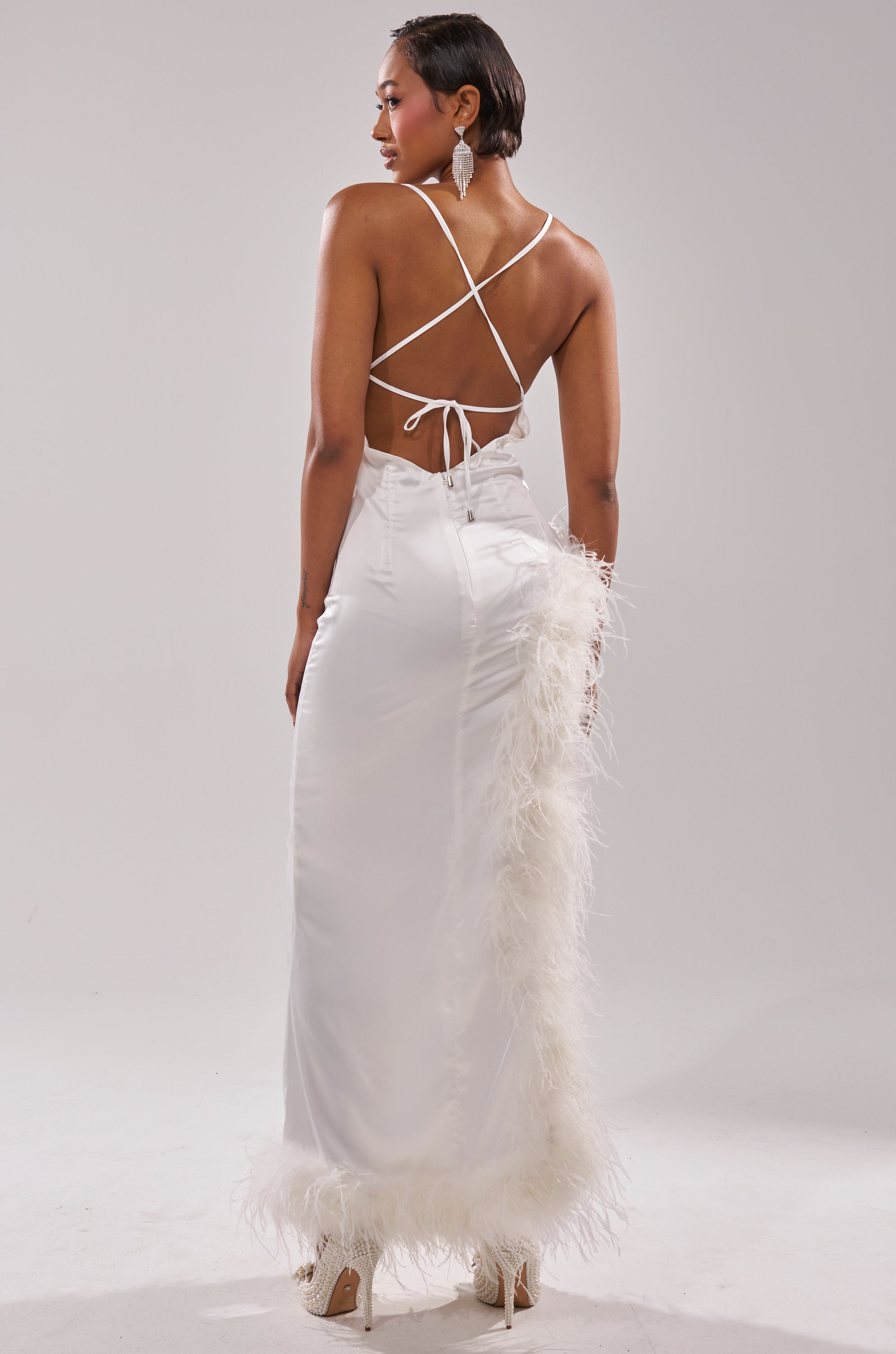 A woman in the LIGHT AS A FEATHER MAXI DRESS—white satin with a crisscross back and feather-trimmed hem and side—faces away against a plain light background, accessorized with heels and statement earrings.