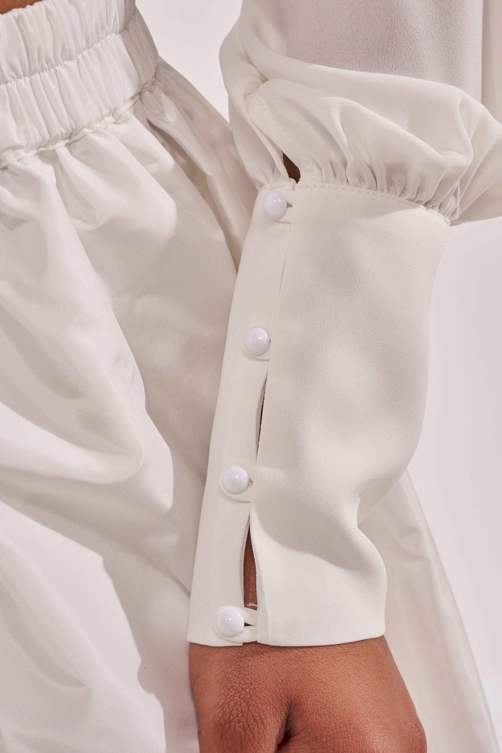 A close-up of the EFFORTLESS CORSET BLOUSE TOP in white, showing long sleeves with three white cuff buttons, an elastic waistband, and soft, slightly shiny fabric. 10